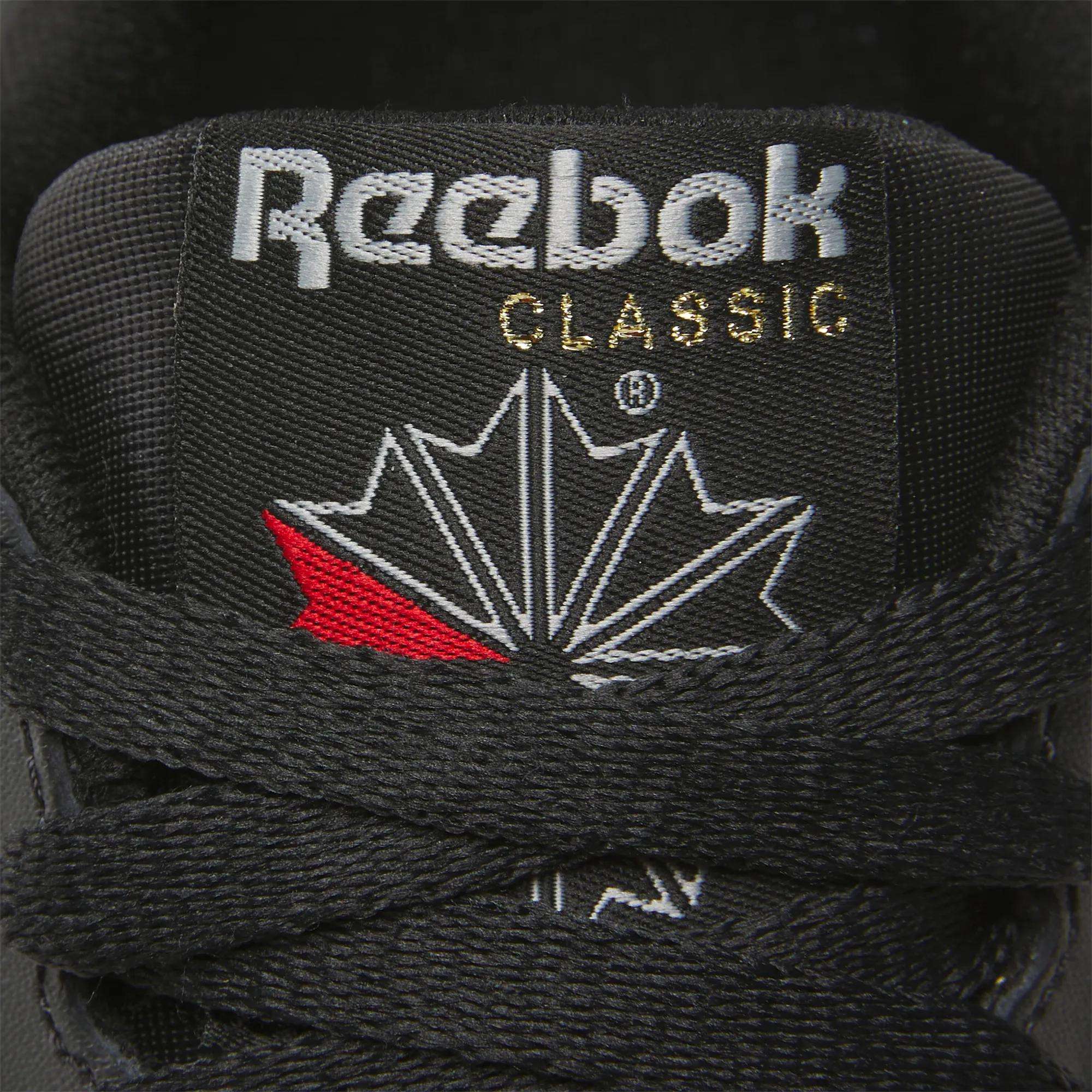 Close-up of the tongue on Women's Classic Leather Shoes by Reebok, featuring the Reebok logo, gold "CLASSIC" text, a white and red geometric maple leaf design above black laces, and a cushioned midsole for comfort.