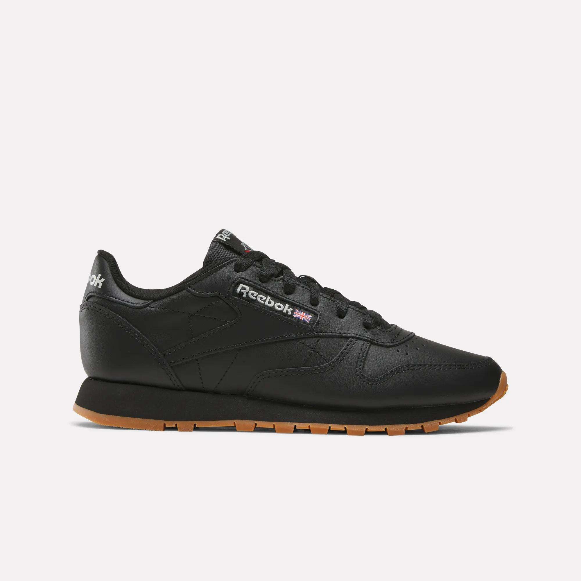 A pair of Women's Classic Leather Shoes by Reebok, featuring a black upper, gum sole, cushioned midsole, black laces, and the Reebok logo on the side, is displayed against a plain white background.