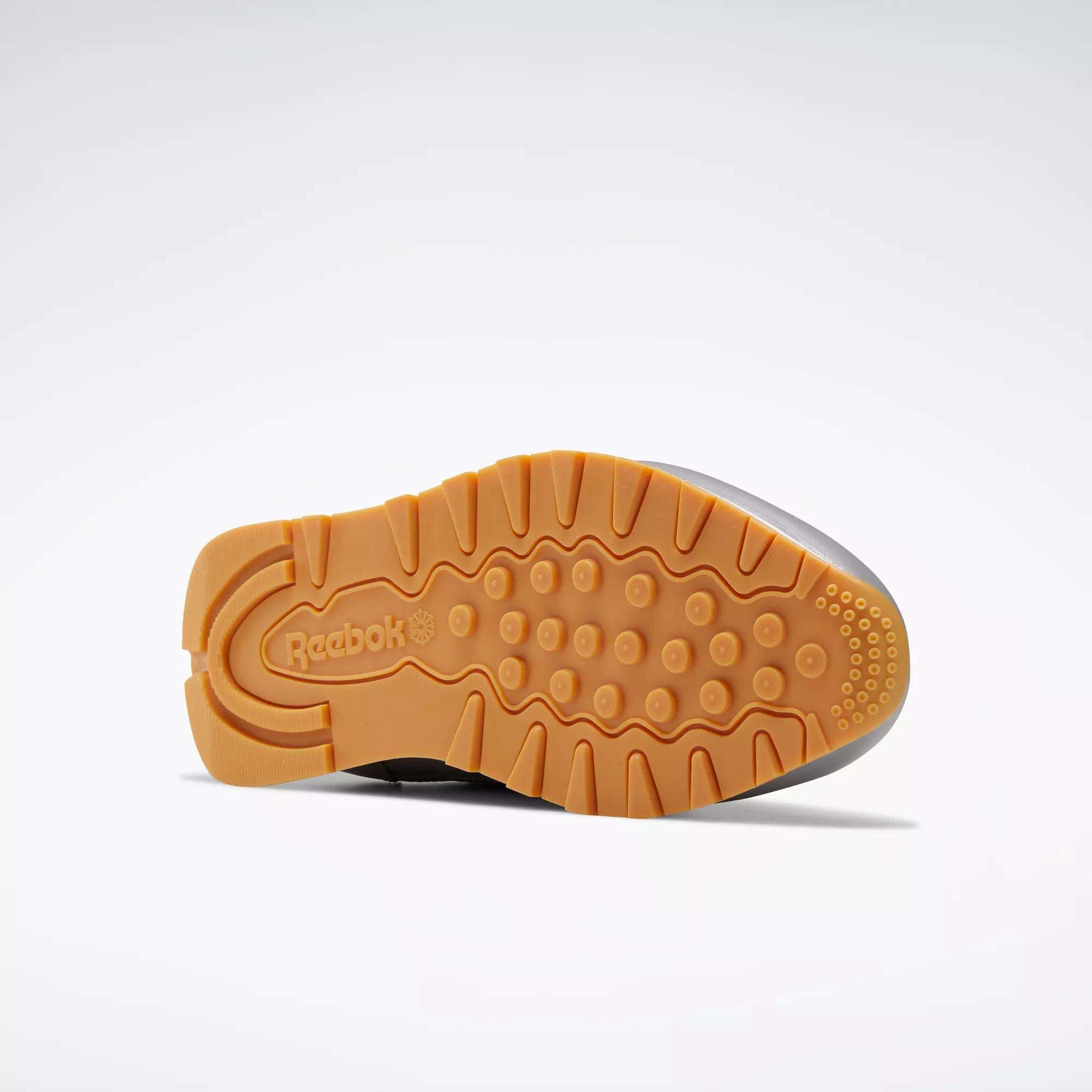 The image displays the bottom of Reebok Classic Leather Shoes, showcasing a gum-colored textured rubber sole with circular and geometric tread patterns for enhanced traction.