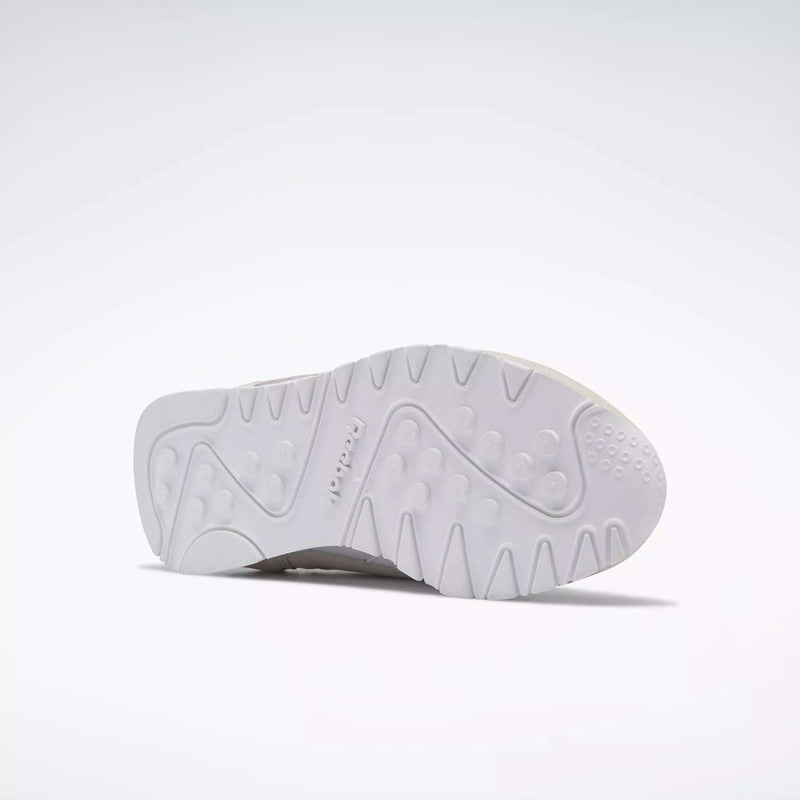 The image shows the white rubber sole of Reebok Women's Classic Nylon Shoes, viewed from below with a textured pattern and circular grooves. The Reebok logo is embossed near the center, emphasizing these versatile everyday sneakers.