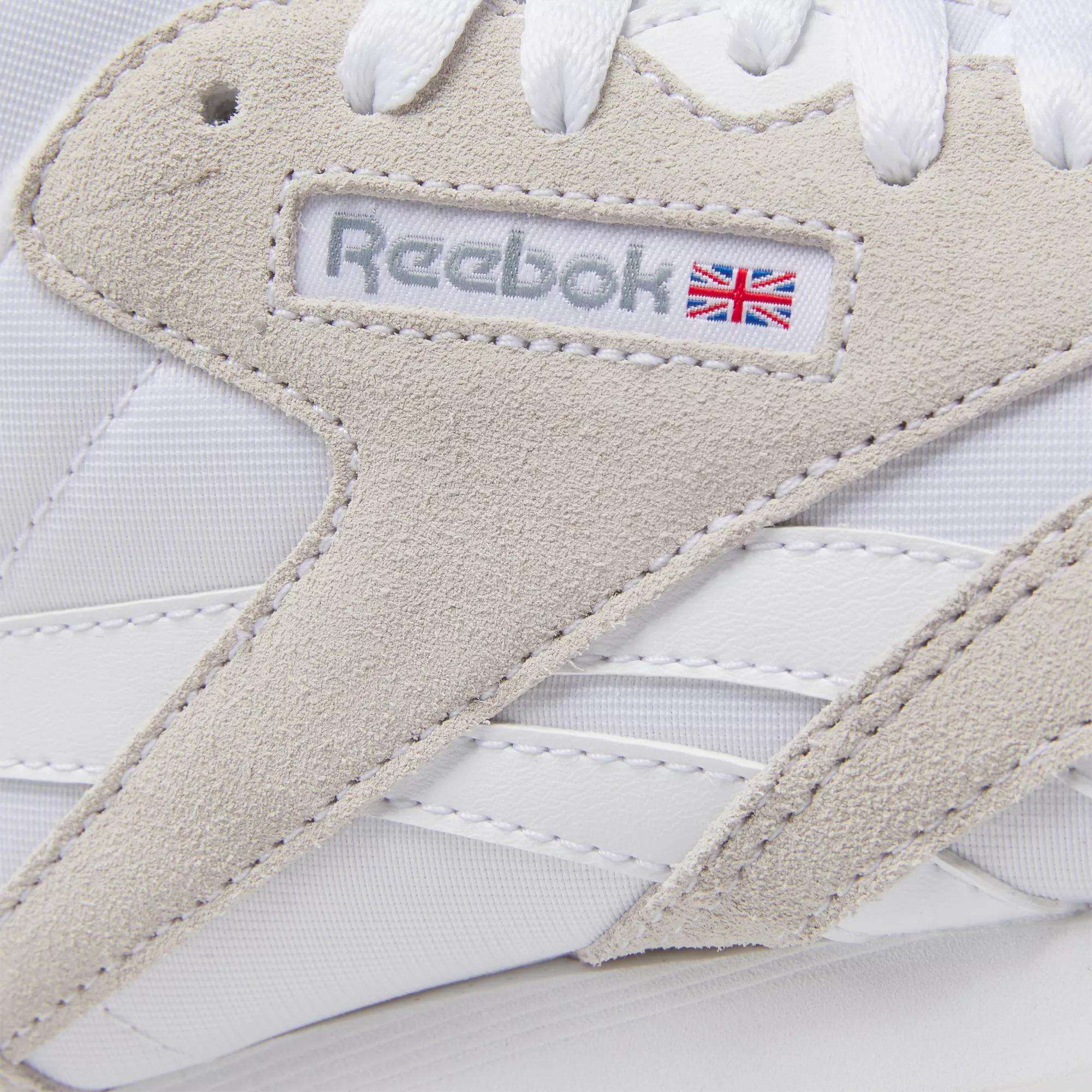 Close-up of Reebok Women's Classic Nylon Shoes in white with beige suede accents, white laces, and the Reebok logo featuring a small UK flag—stylish everyday sneakers.