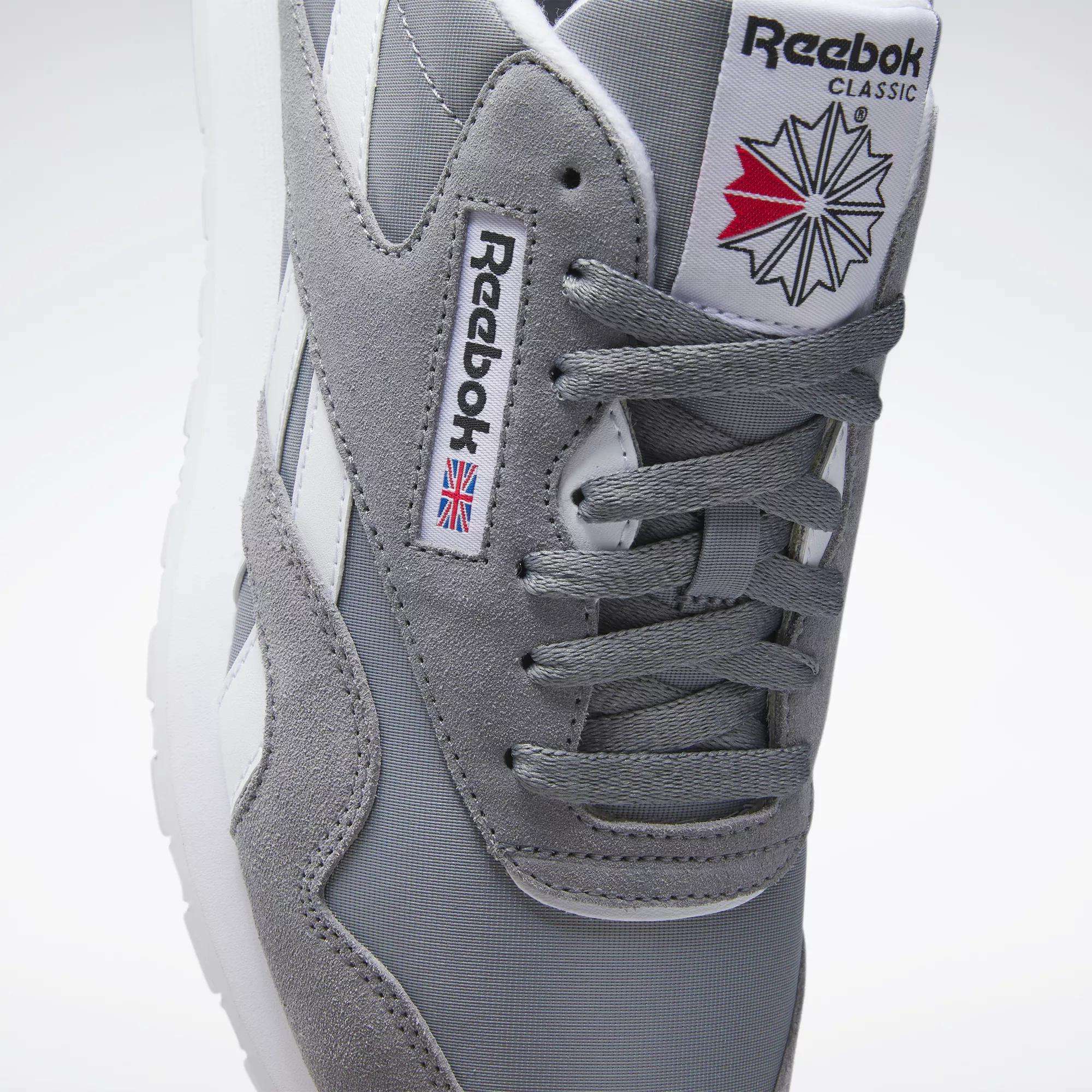 A close-up of Reebok Men's Classic Nylon Shoes with a grey nylon upper, white stripes, laces, Reebok branding, and a red, white, and blue geometric design on the tongue for classic everyday style.