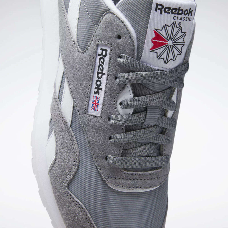 A close-up of Reebok Men's Classic Nylon Shoes with a grey nylon upper, white stripes, laces, Reebok branding, and a red, white, and blue geometric design on the tongue for classic everyday style.