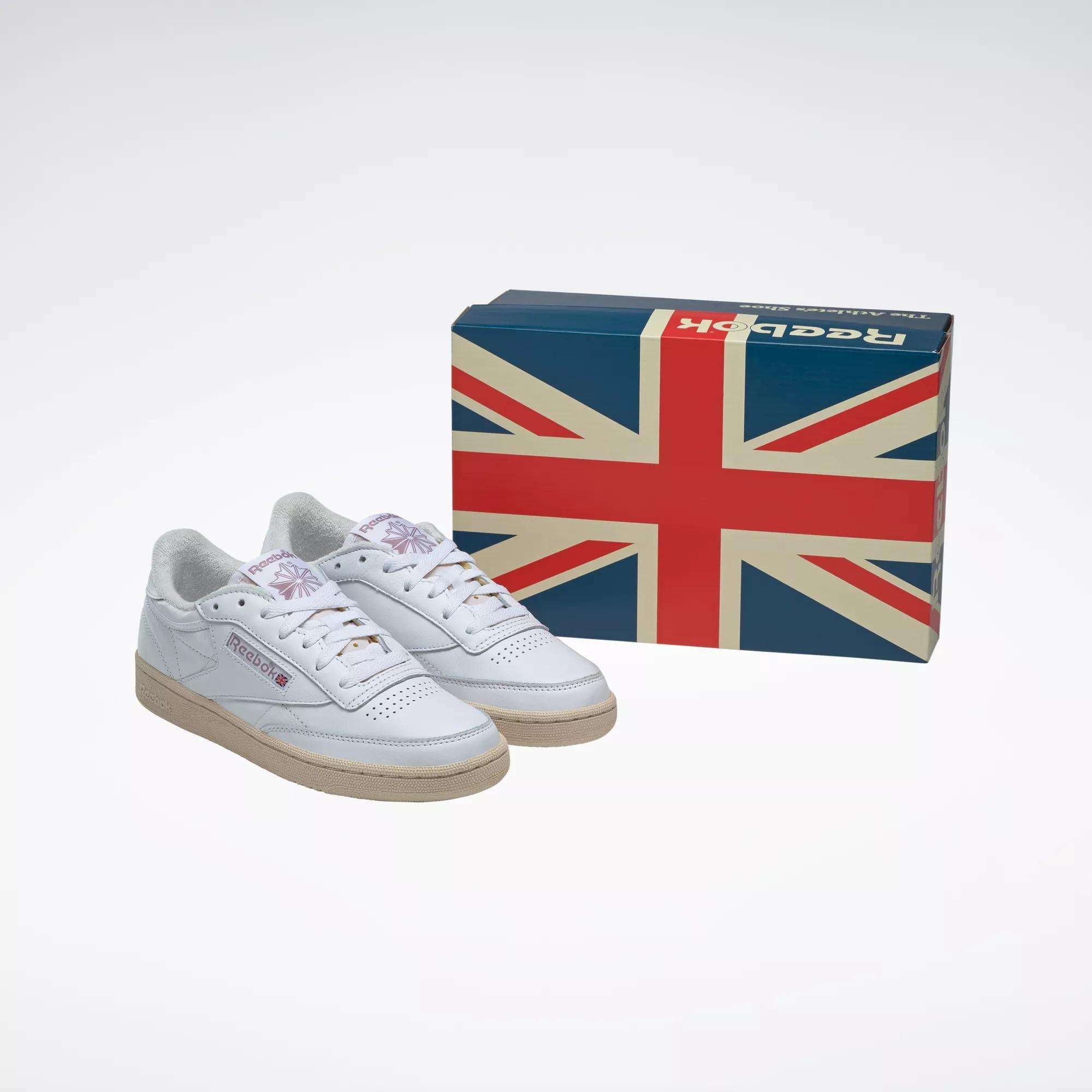 A pair of Women's Club C 85 Vintage Shoes by Reebok, in white with gum soles, are placed next to a shoebox featuring a large Union Jack flag against a plain background.