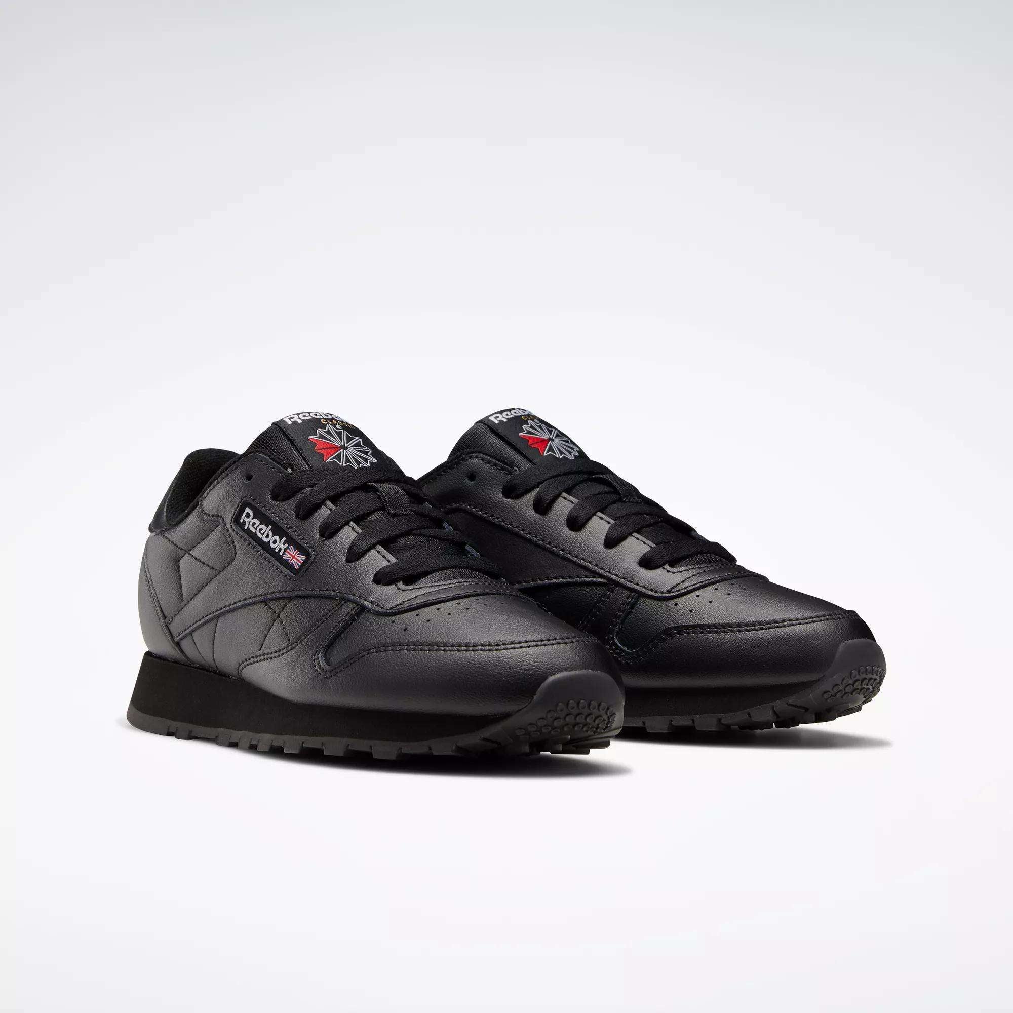 A pair of Classic Leather Shoes - Big Kids by Reebok features black leather, black laces, black soles, and the Reebok logo on the side and tongue, shown against a light gray background.