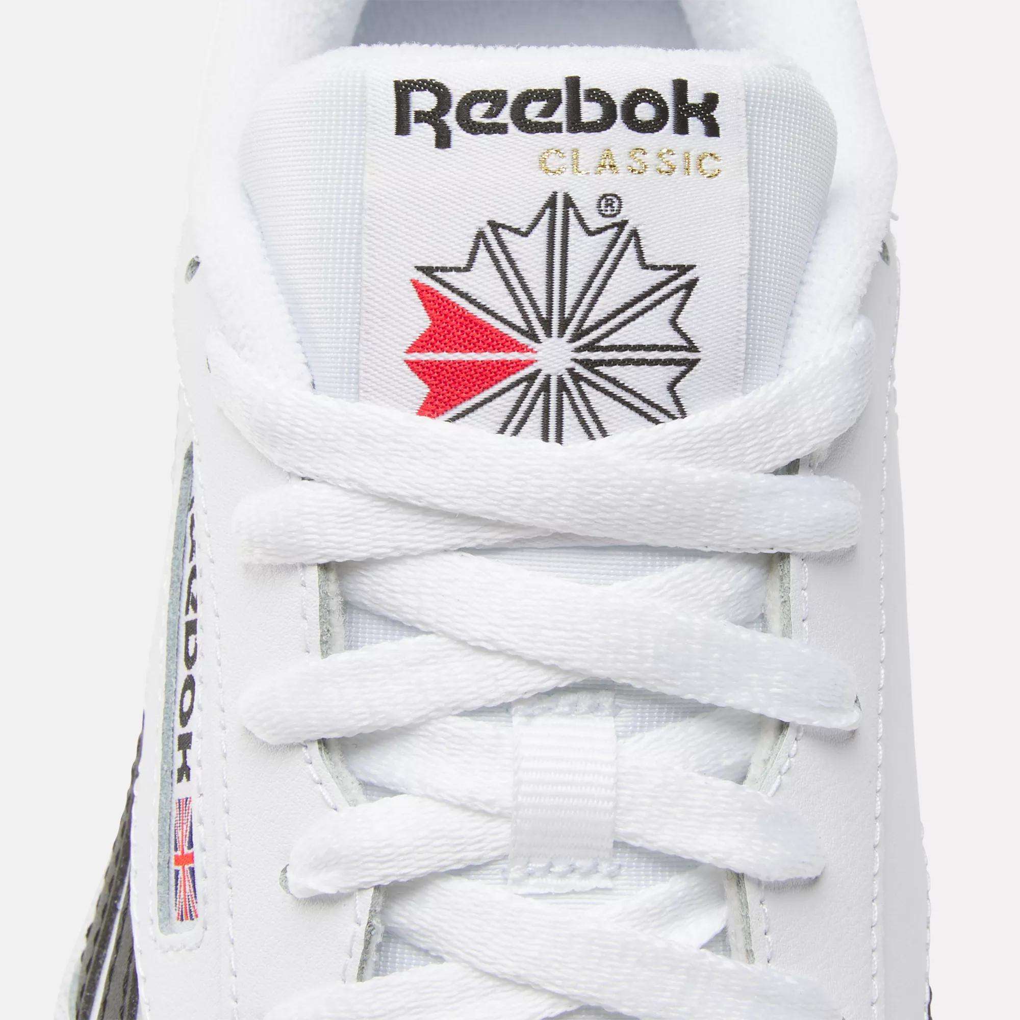 Close-up of a white Reebok Club C Revenge shoe displaying the tongue with the Reebok logo, red and black star design, and white laces. The Reebok name and small UK flag are visible on these casual leather shoes.
