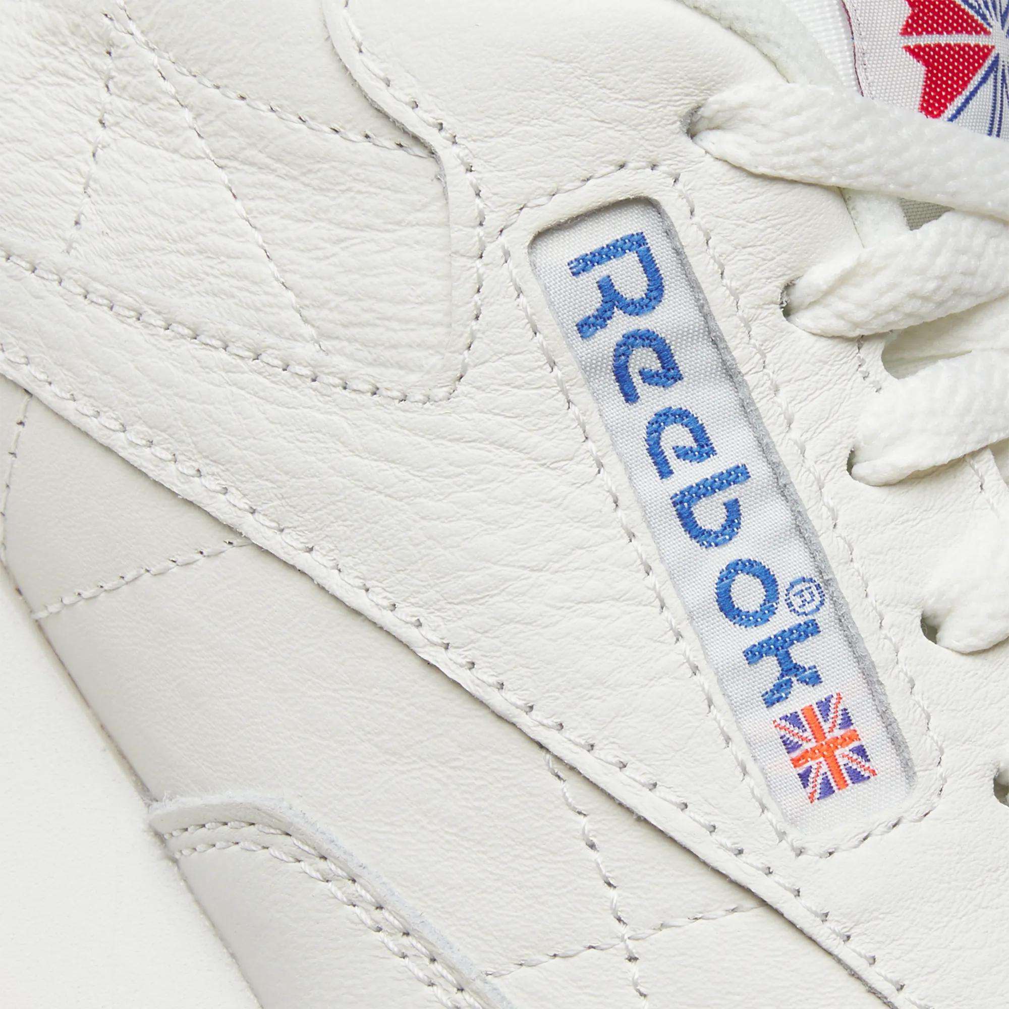 Close-up of Reebok Classic Leather Shoes shows the blue Reebok logo and a small Union Jack on the durable white leather side panel, with white laces highlighting their retro style.