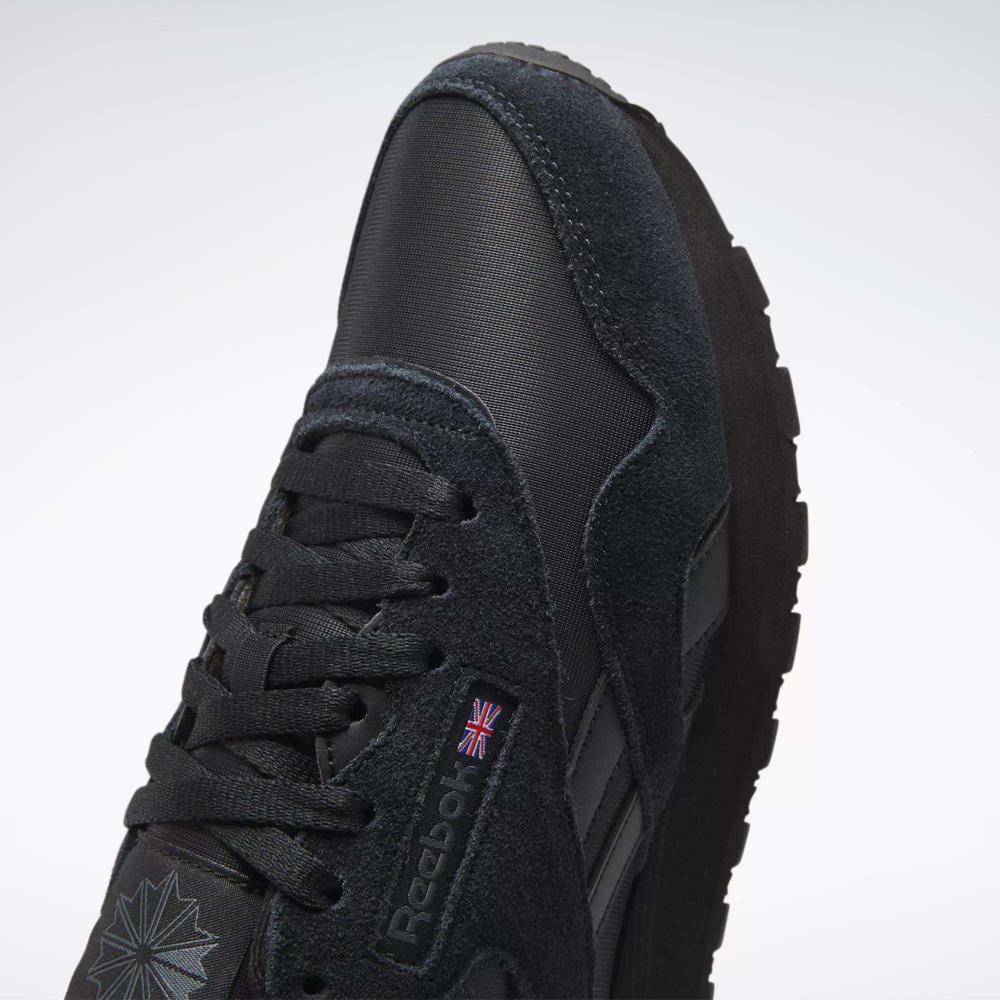 Close-up of the Reebok Men's Classic Nylon Shoes in black, featuring a suede and nylon upper, laces, Reebok logo with UK flag, thick rubber sole, and a star-shaped design near the toe for a classic everyday look.