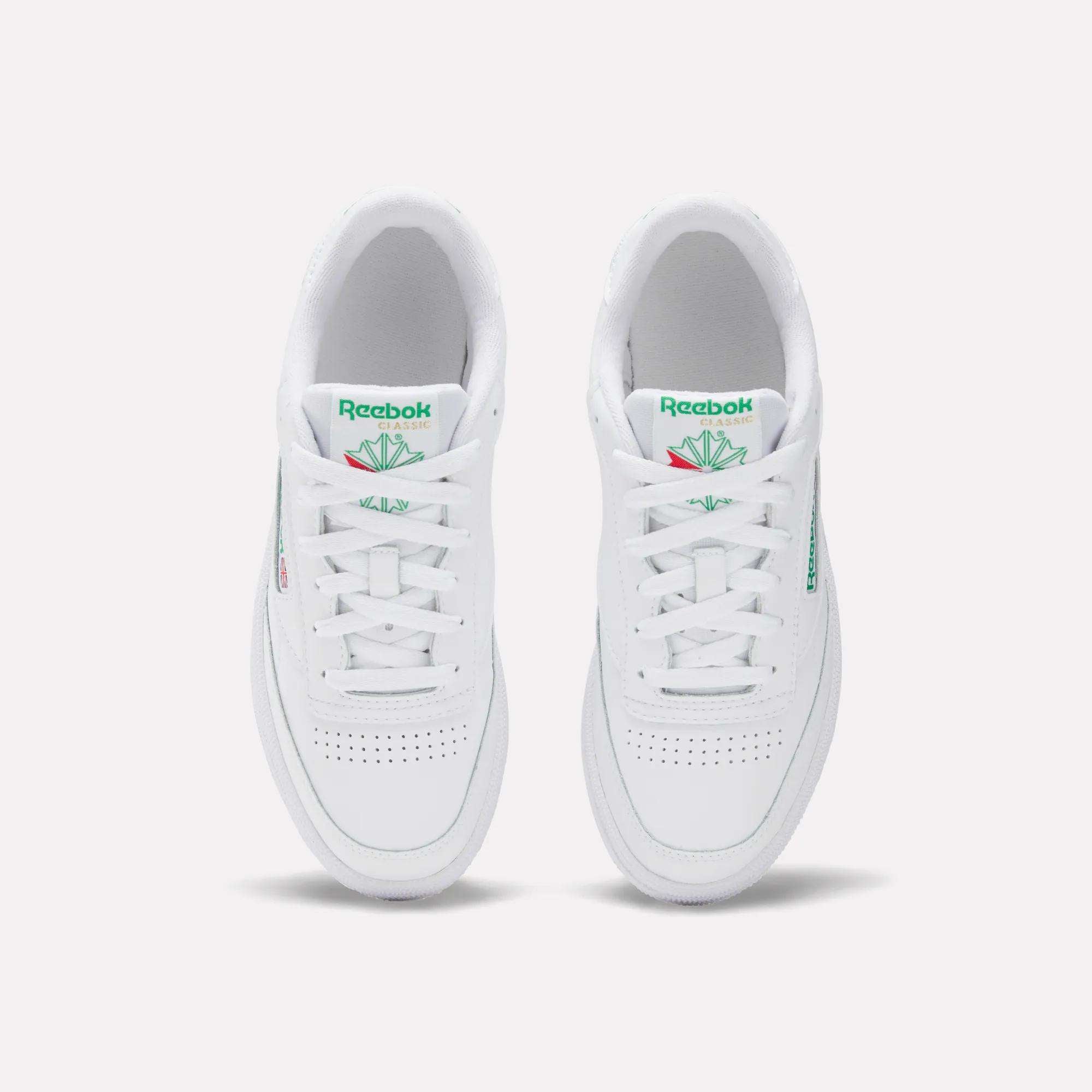 The Women's Club C 85 Shoes by Reebok are white sneakers with green Reebok logos and tongue detailing, a clean minimalist look, and toe perforations, shown from an overhead view.