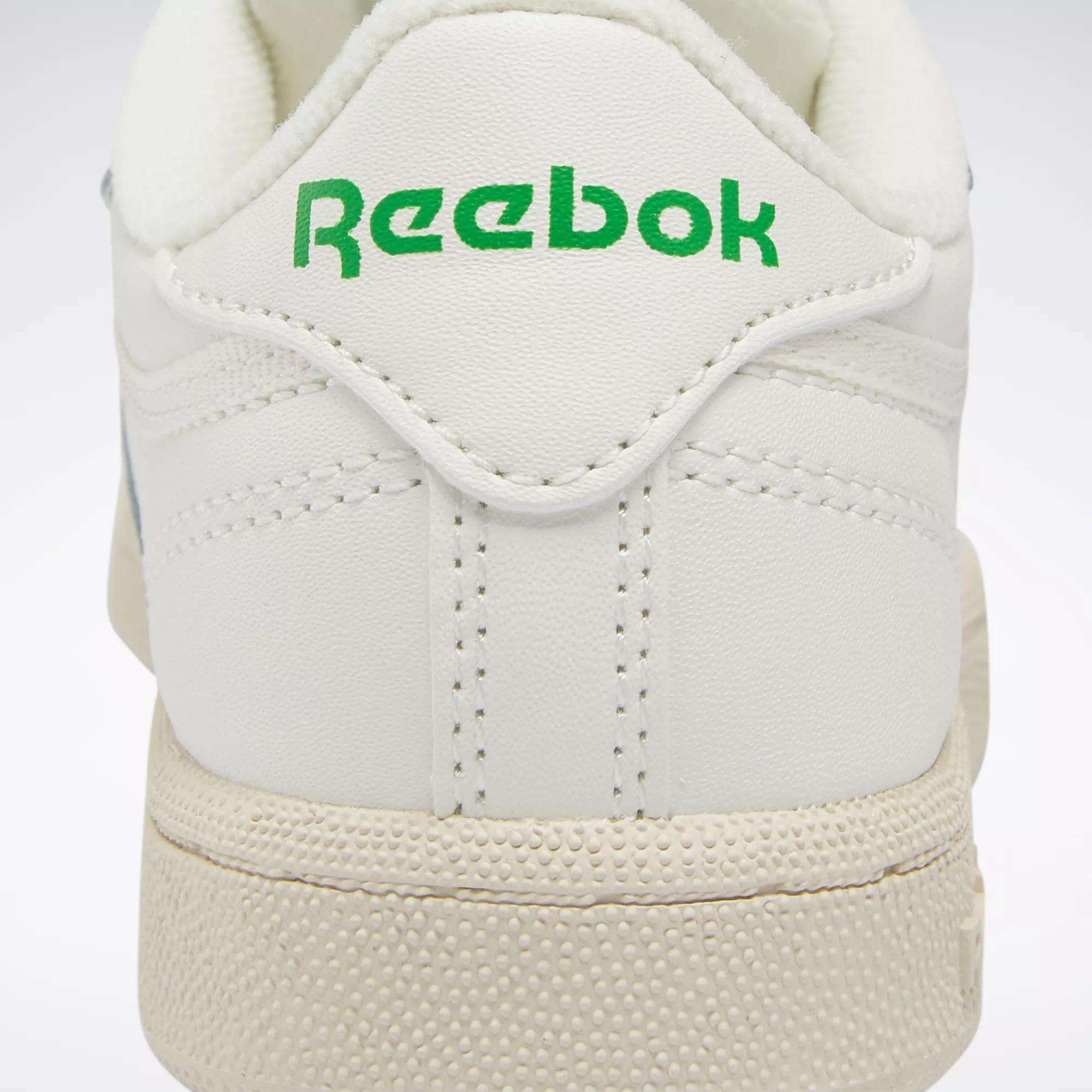 Close-up of the back of a white Reebok Kids' Club C Shoe for preschoolers, featuring "Reebok" in green on the heel tab, textured off-white soles, and detailed stitching inspired by '80s style.