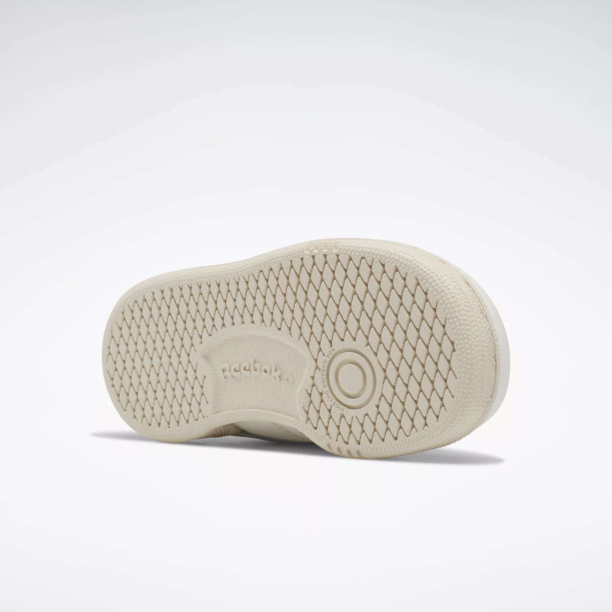 The image shows the beige sole of Reebok Kids' Club C Shoes for babies and toddlers, featuring a textured diamond pattern, circular details, and an embossed Reebok logo at the center, all set against a light background.
