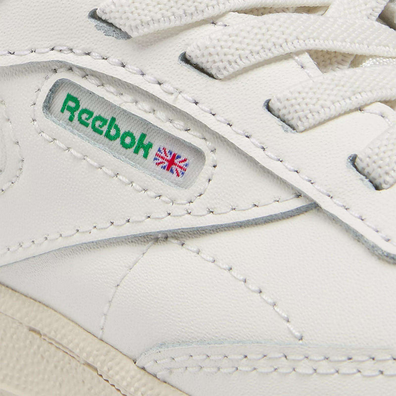 Close-up of Reebok Kids' Club C Shoes for baby & toddler in classic '80s style, featuring the green Reebok logo, small Union Jack flag on the side, visible stitching, and white laces.