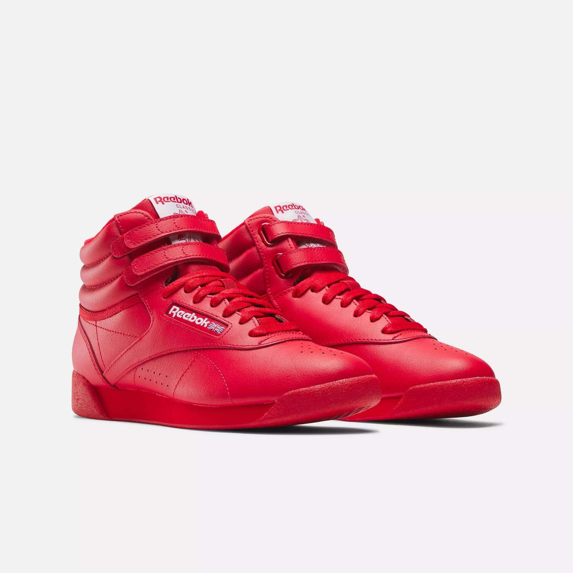 The Reebok Women's Freestyle Hi Shoes are bright red high tops with velcro straps and laces, shown on a white background. The Reebok logo appears on the side and tongue, making them ideal women's fitness shoes.