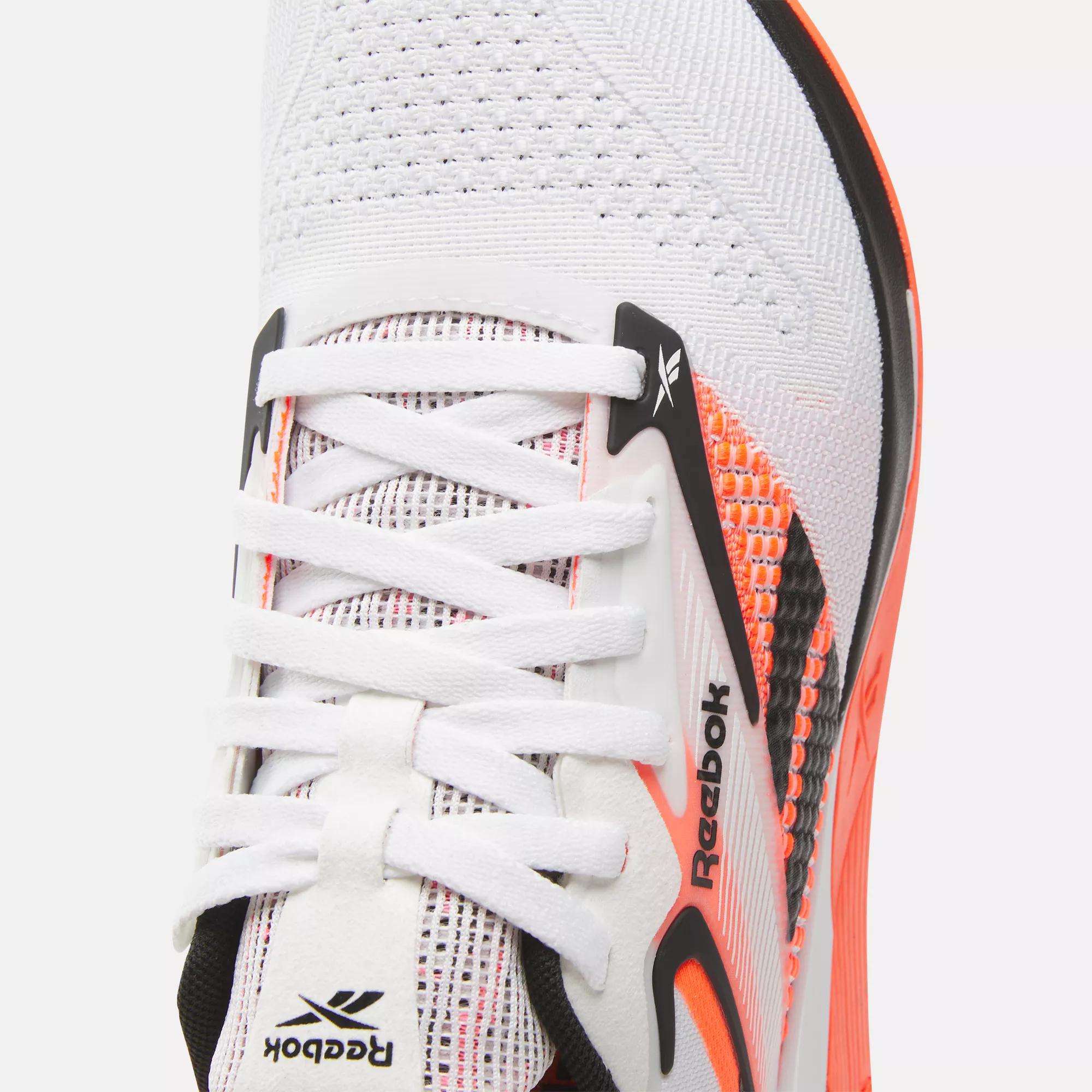 Close-up of the Reebok Women's Nano X4 Training Shoes in white with black and orange accents, featuring breathable mesh upper, laces, and Reebok logo near the eyelets—perfect lightweight option for daily workouts.
