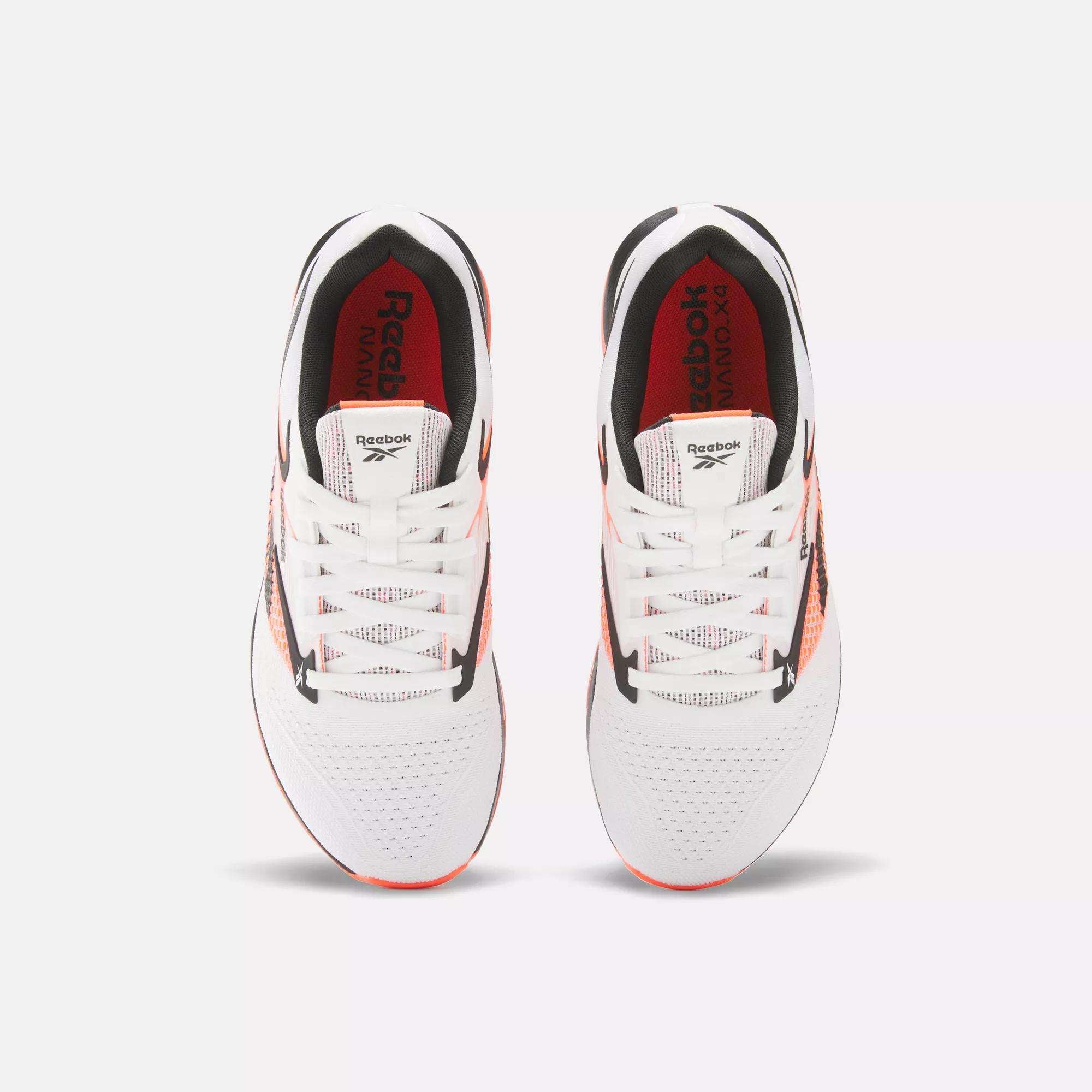 Top view of a pair of Reebok Women's Nano X4 Training Shoes in white with black and orange accents, red insoles, and white laces, side by side on a light background. These lightweight shoes are designed for peak performance.