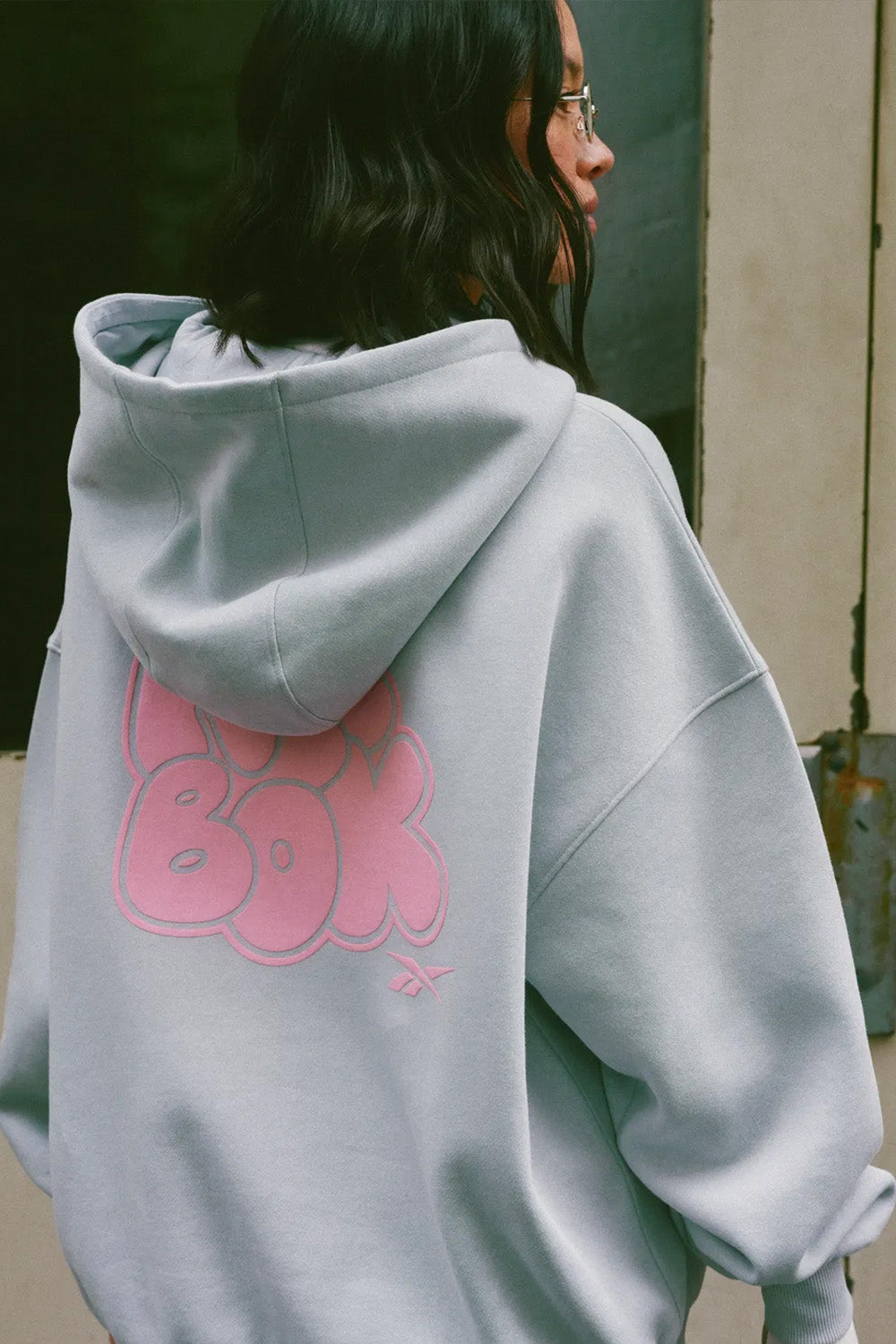 A person shown from the back wearing an oversized light gray hoodie with a large pink 'BOY' graphic printed on the back, photographed in an outdoor urban setting.