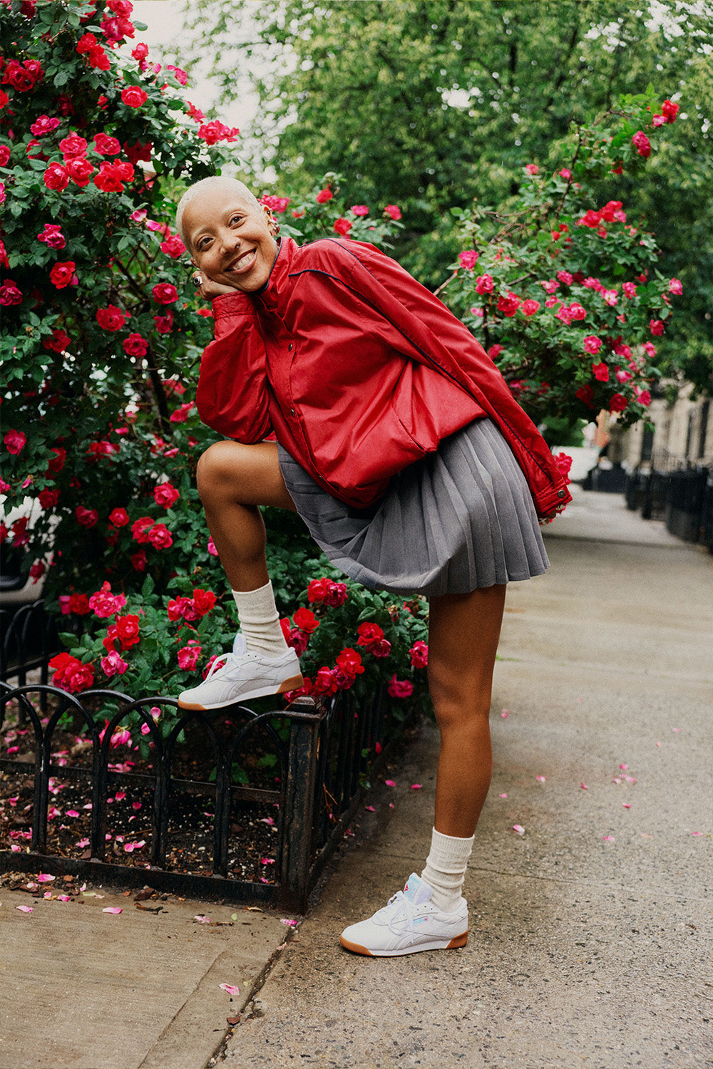 woman wearing red shirt and gray skirt  and white sneakers, smiling against a rose bush 