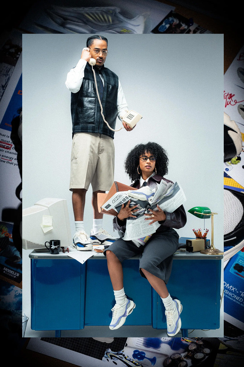 A fashion editorial-style photo featuring two models in a retro office scene. One model stands holding a vintage telephone, wearing a black leather vest over a white long-sleeve shirt, beige shorts, and white sneakers.