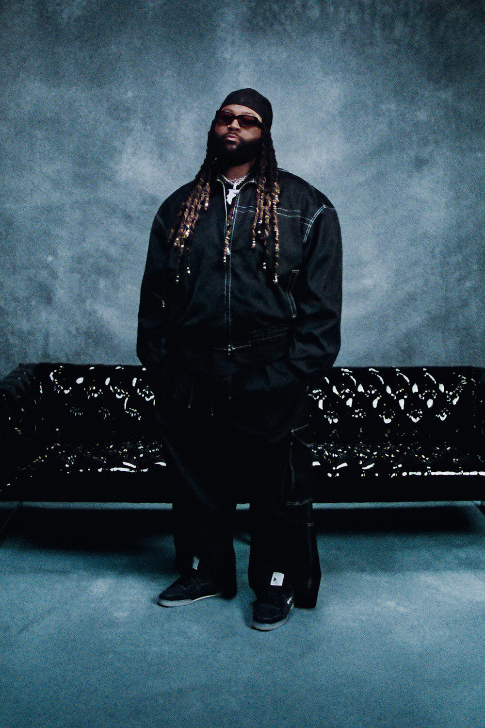 Person wearing a black outfit with long braids sitting on a black couch against a textured gray wall.