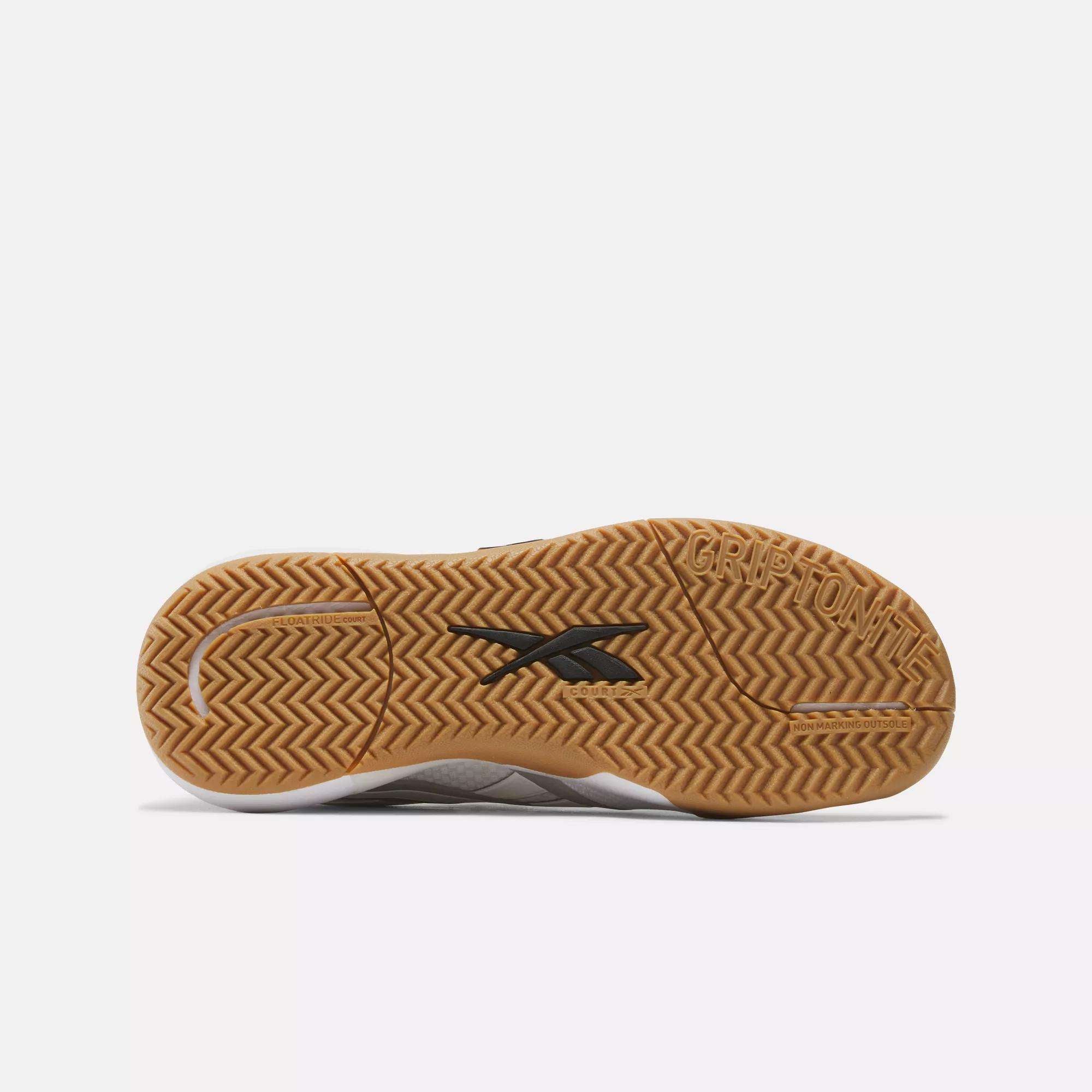 The image shows the sole of a Women's Nano Court Training Shoe by Reebok, with a brown gum-rubber bottom, herringbone tread for traction, a black Reebok logo in the center, and "GRIPTONITE" near the heel.