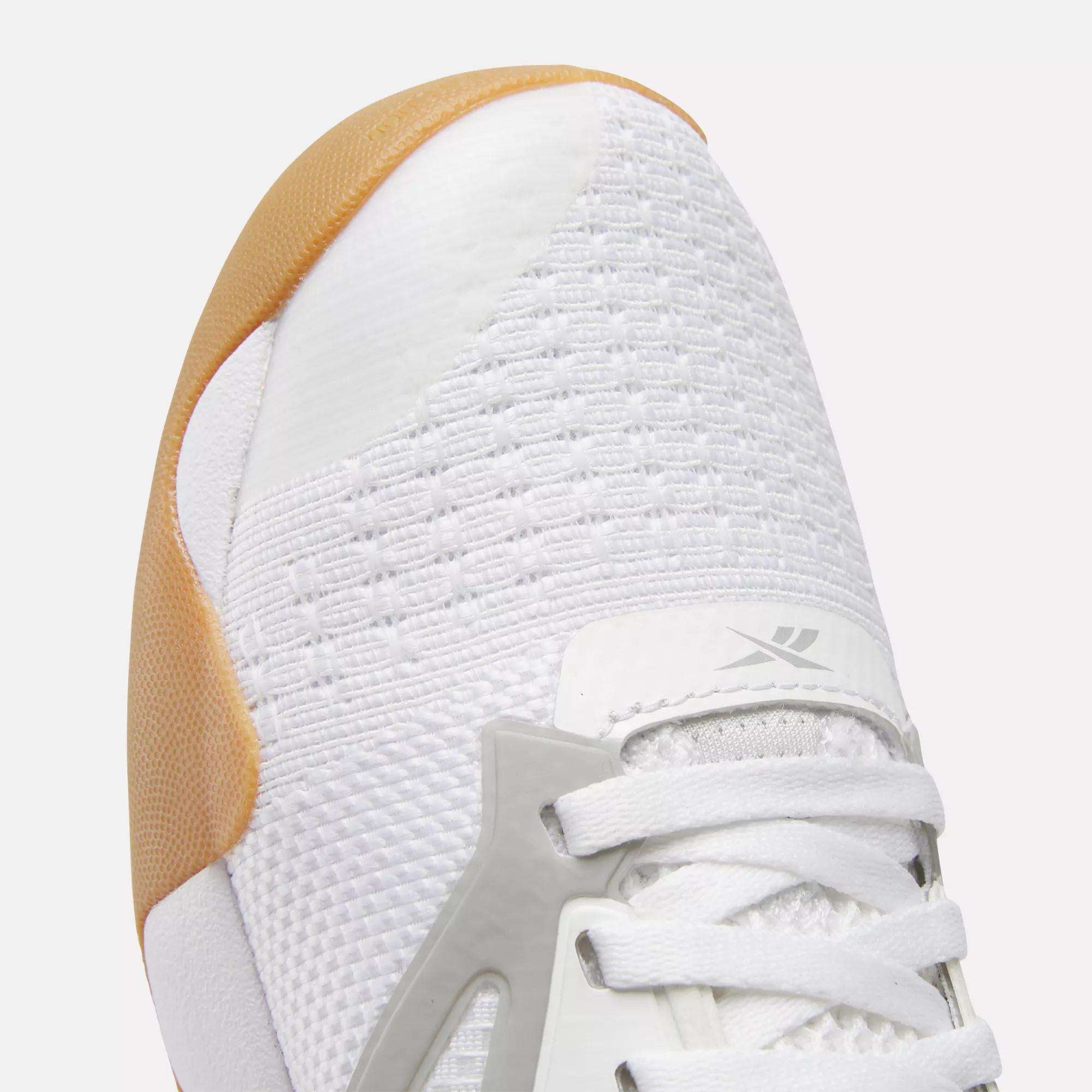 Close-up of the front of Reebok Women's Nano Court Training Shoes, featuring a light brown rubber toe cap, breathable mesh, white laces, and a silver Reebok logo on the tongue—designed for multidirectional traction.