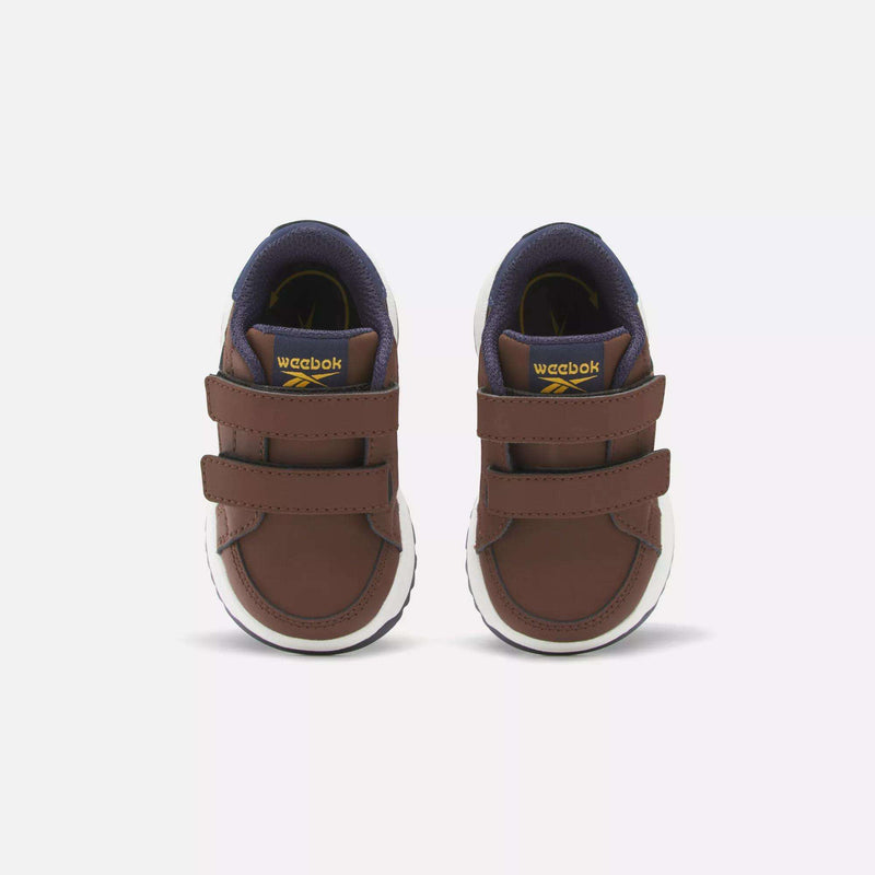 The Reebok Weebok Clasp Low Shoes for infants and toddlers are brown with white soles, double Velcro straps, a dark blue inner lining, and yellow tongue accents, shown facing forward on a plain light background.