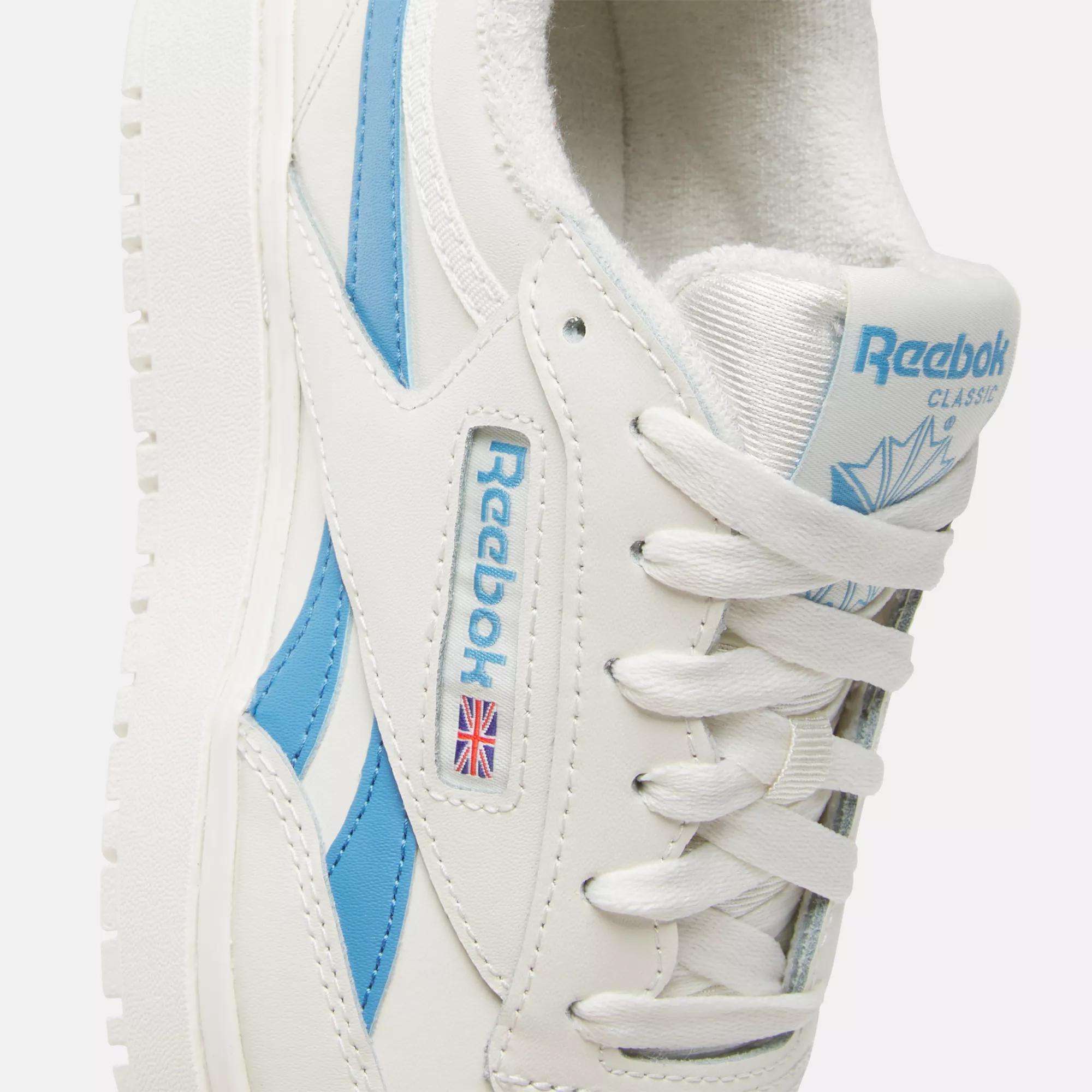Close-up of Reebok Women's Club C Double Revenge Shoes featuring a white leather upper, light blue accents, white laces, and the Reebok logo with a small Union Jack on the side. The tongue displays blue Reebok branding.