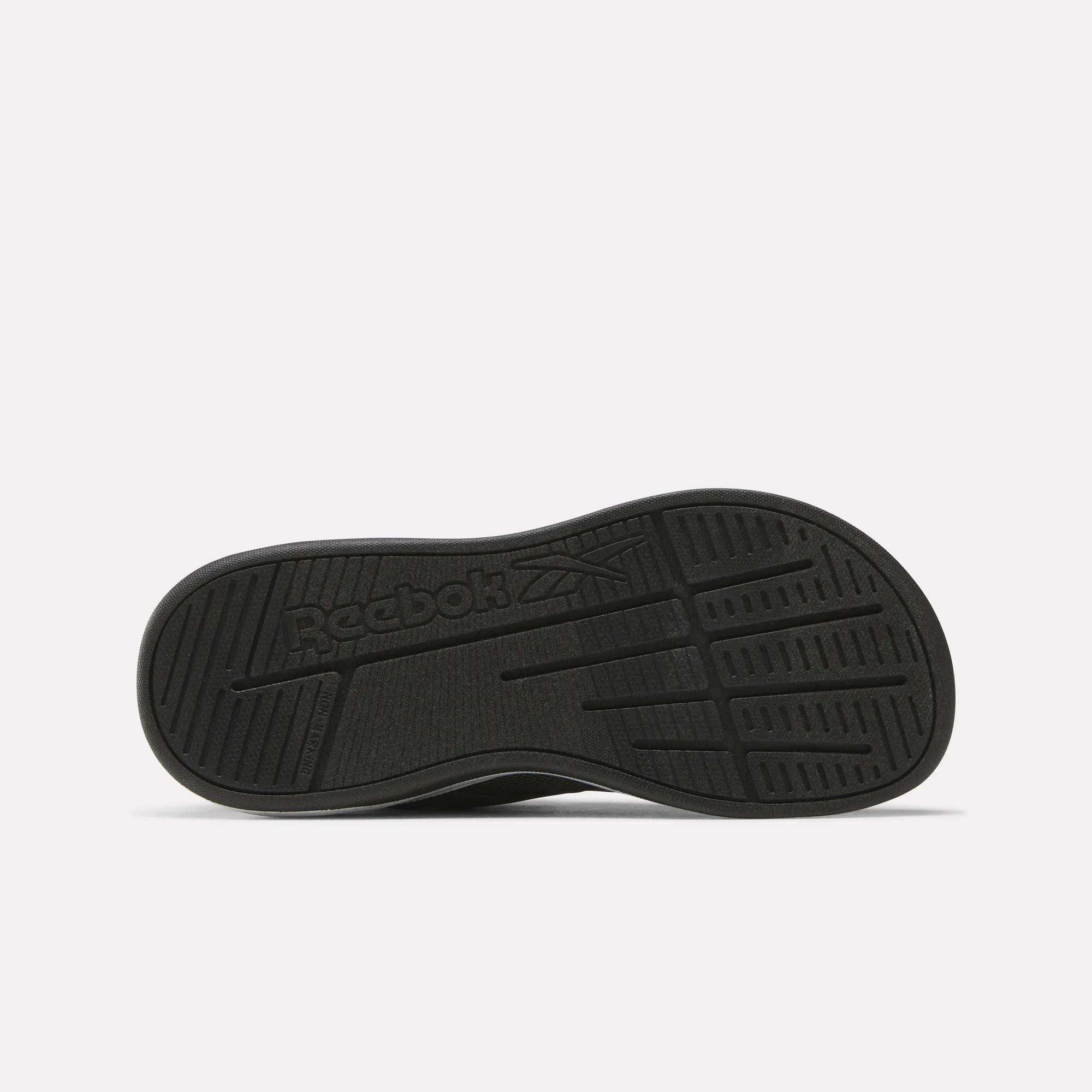 The image shows the black rubber outsole of the Reebok Nano Play Stretch Lace And Top Strap Shoes for little kids, with textured grip patterns and an embossed Reebok logo featuring the brand's vector symbol in the center.