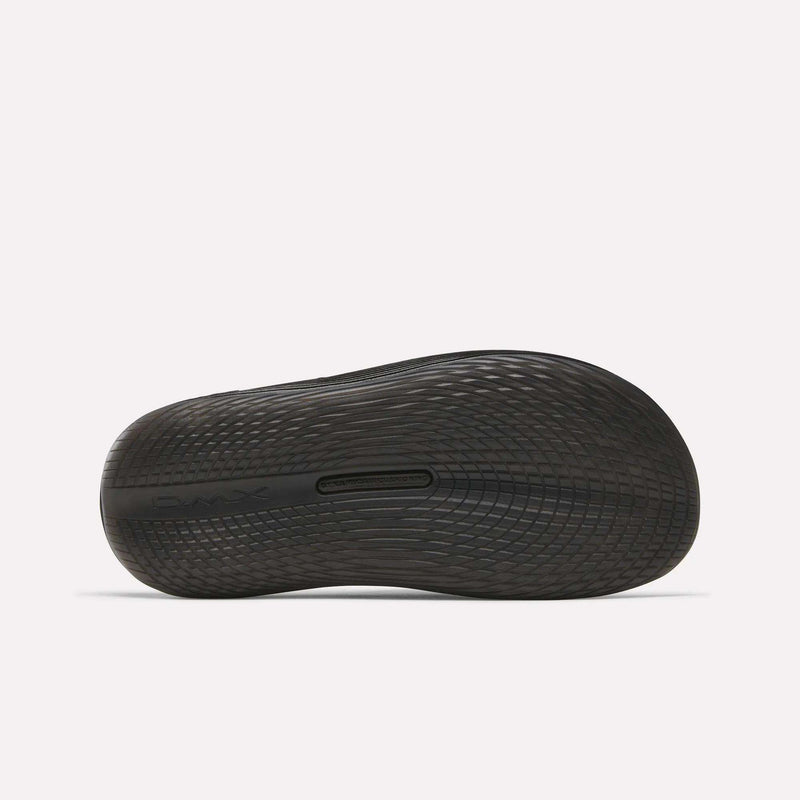 The image shows the black, textured sole of a Reebok DMX Recovery Walking Slide viewed from the bottom, highlighting its tread pattern and cushioning, with the Reebok logo embossed near the heel.