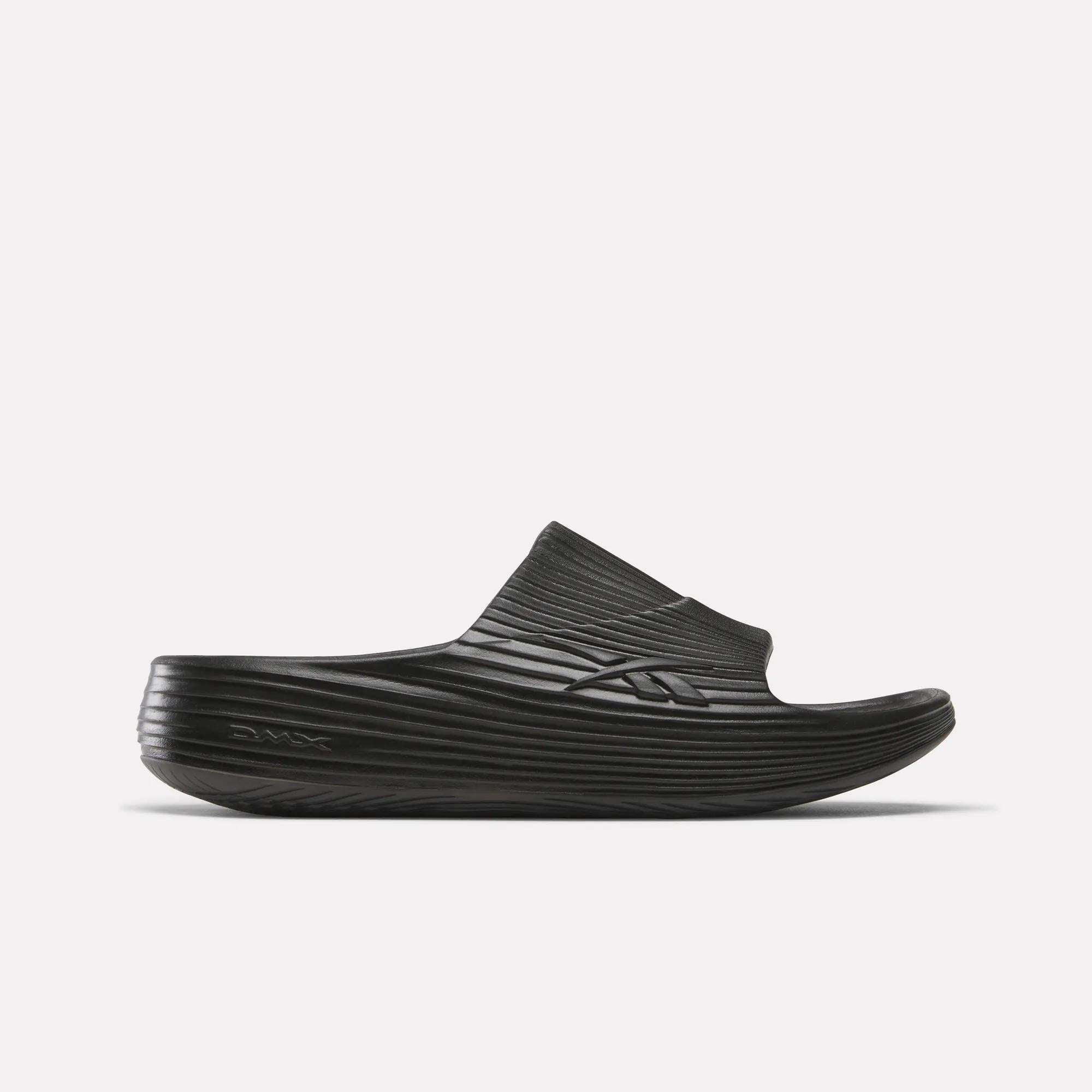 A single Reebok DMX Recovery Walking Slide in black, featuring a textured ridged design, thick sole, super-soft foam, and open toe, is displayed against a plain white background.