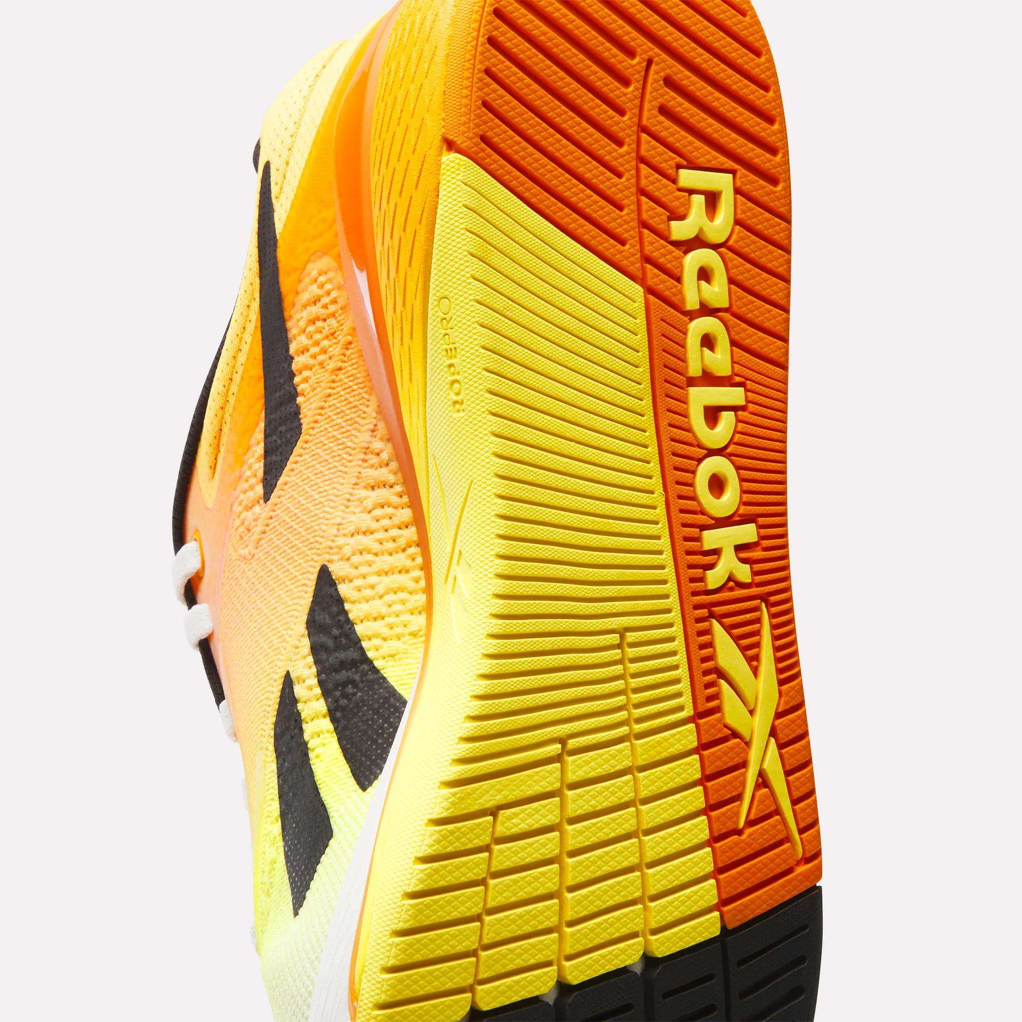 Close-up view of the Nano X5 Training Shoes by Reebok, showing the bright yellow, orange, and black sole with the Reebok logo and a large X design on the bottom.
