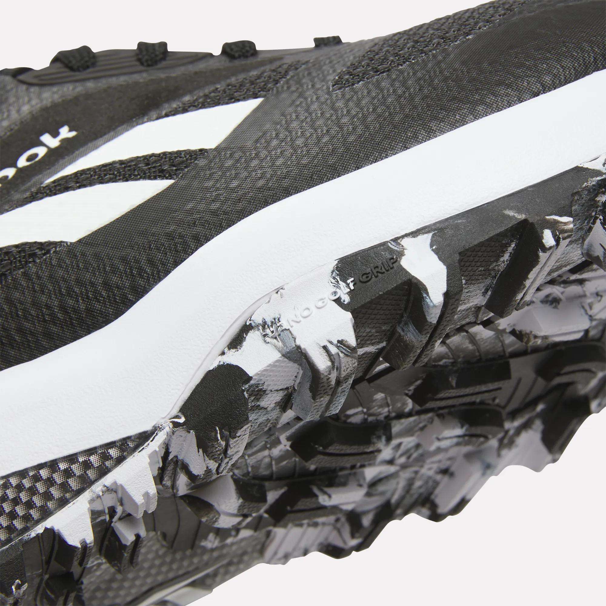 Close-up of Reebok Nano Golf Shoes in black and white, highlighting the textured sole with rugged tread pattern and FlexWeave upper for enhanced durability, plus a visible white logo.