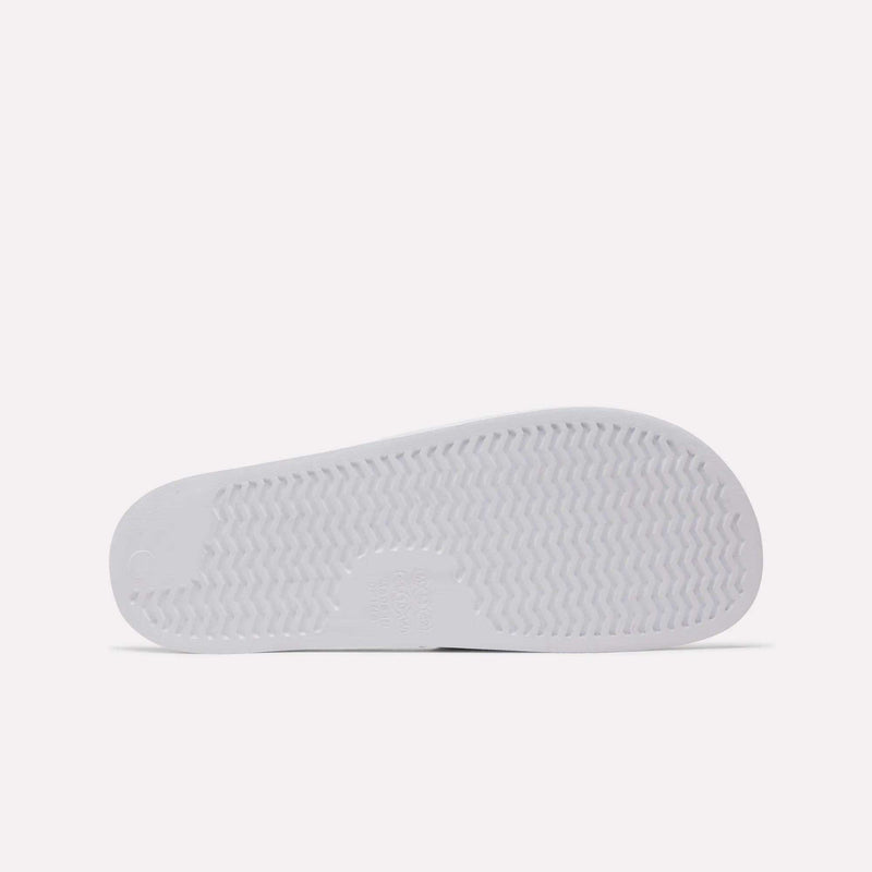 The image shows the bottom of a white Reebok RBK Fulgere Swim Slides sole with a zigzag tread for grip, a smooth circular heel, and a rectangular center section. The plain white background highlights the design.