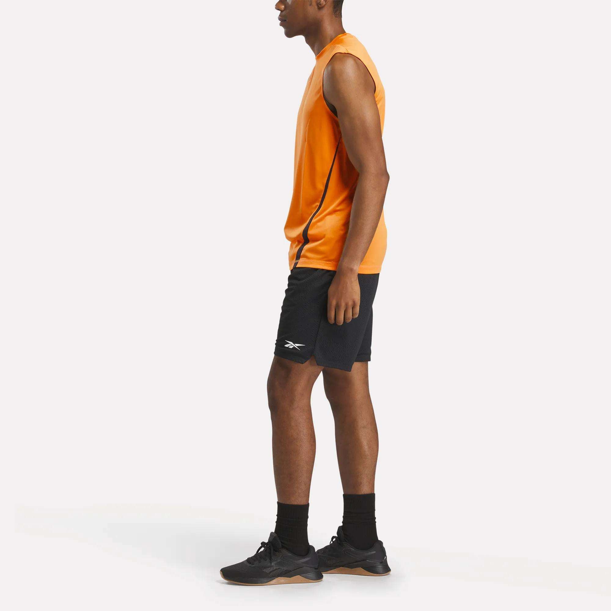 A man in profile wears a Reebok Men's Training Sleeveless Tech T-Shirt in orange, paired with black shorts, socks, and athletic shoes, standing against a plain white background.