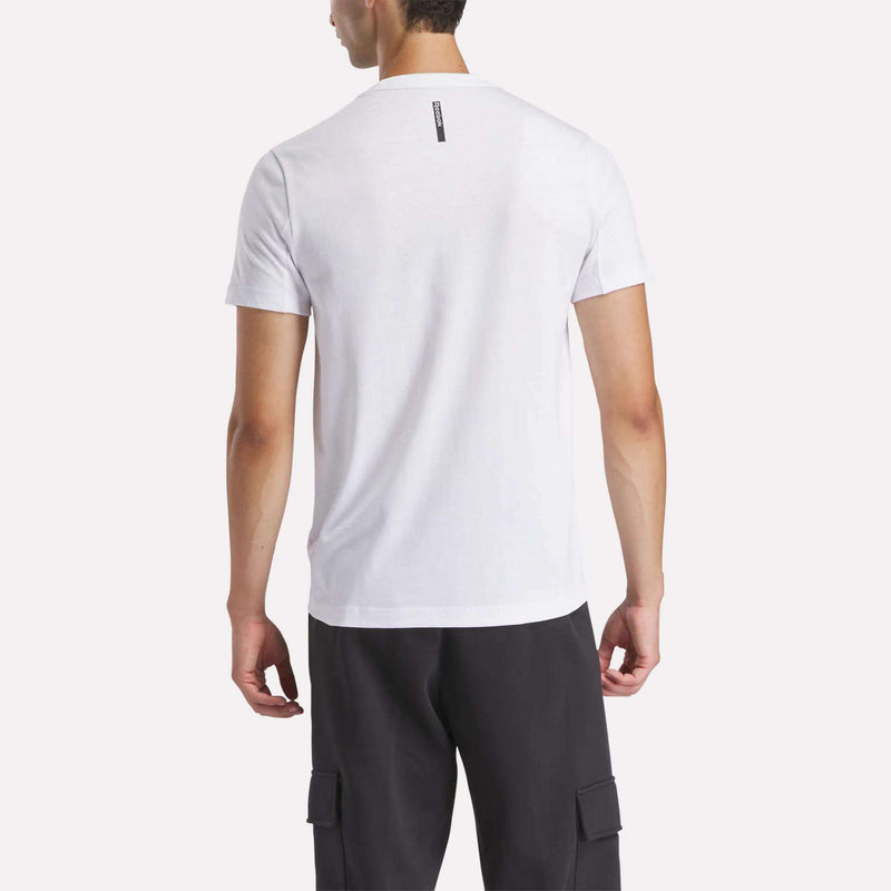 A man is seen from the back wearing the Reebok Men's Athlete Training T-Shirt in white with an athletic fit, paired with black cargo pants, standing against a light background.