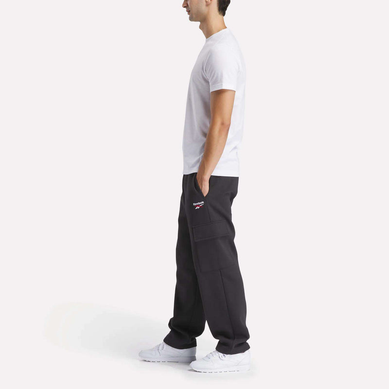 A man stands in profile against a plain background, wearing the Reebok Men's Athlete Training T-Shirt, black cargo pants with his hands in the pockets, and white sneakers.