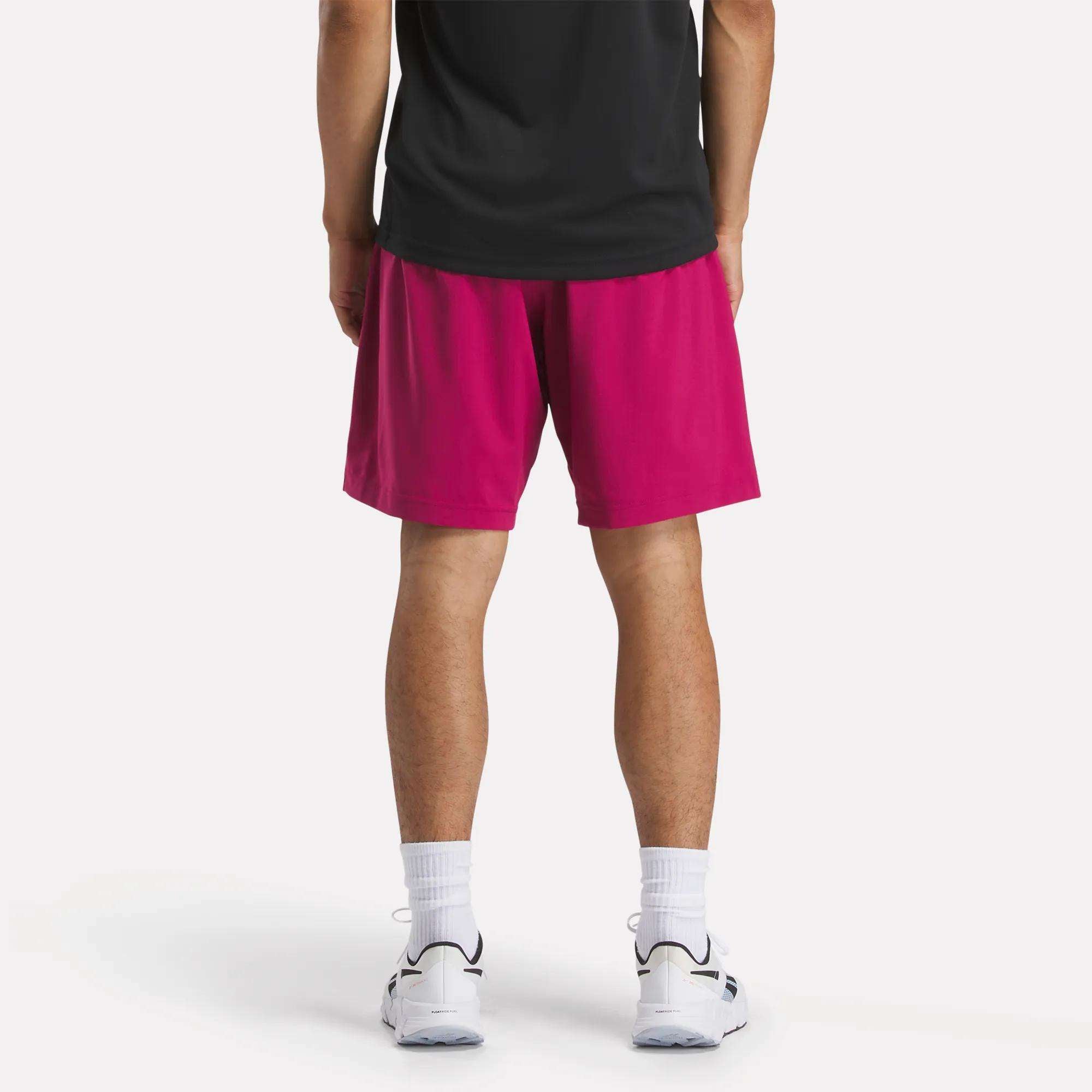 A person is seen from behind wearing a black shirt, Reebok Men's Workout Ready Shorts 9" in bright pink, white crew socks, and white athletic shoes, all set against a plain light background.