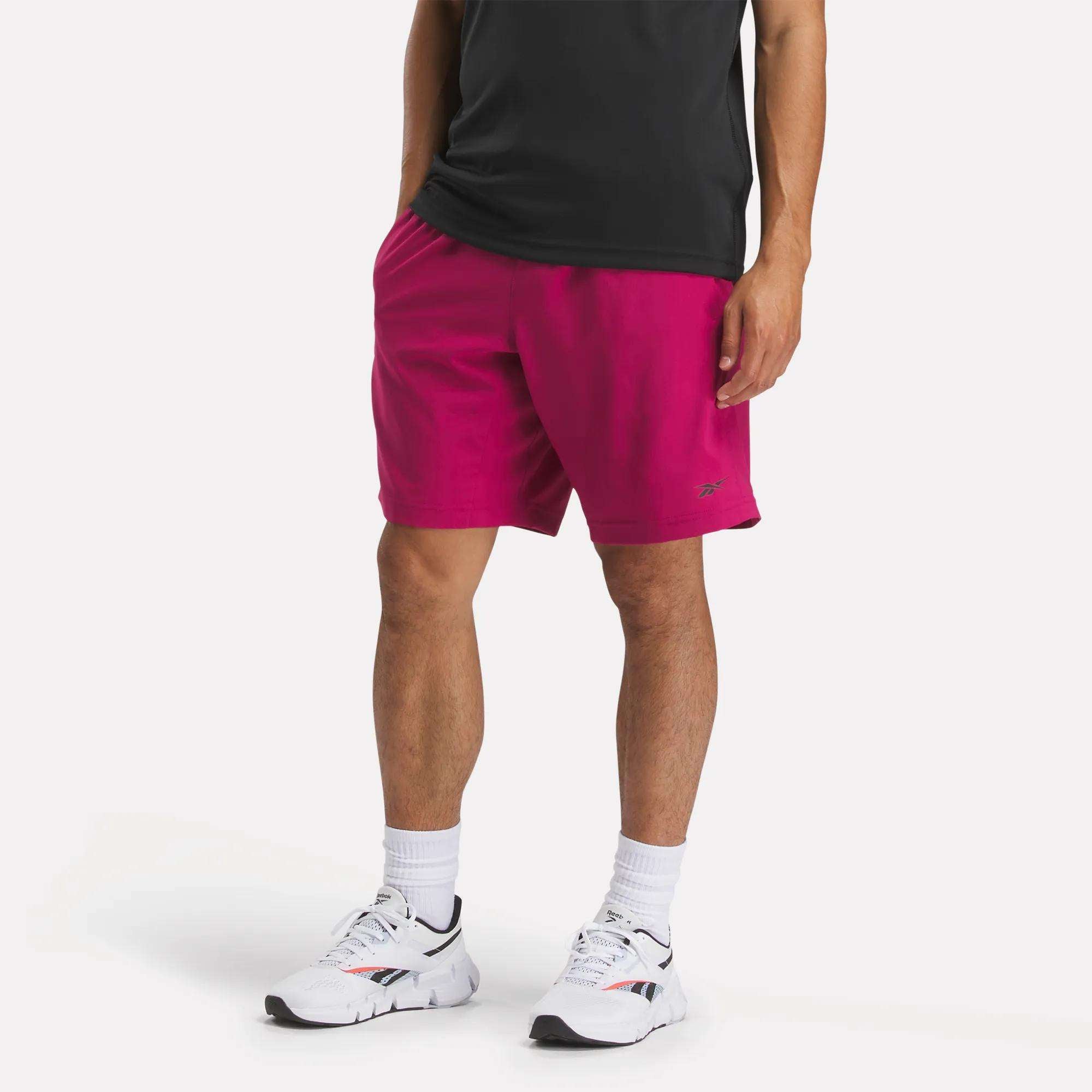 A man is seen wearing a black t-shirt, white crew socks, white sneakers with black and red details, and bright pink Reebok Men's Workout Ready Shorts 9" made with Speedwick fabric. His face is not visible in the image.