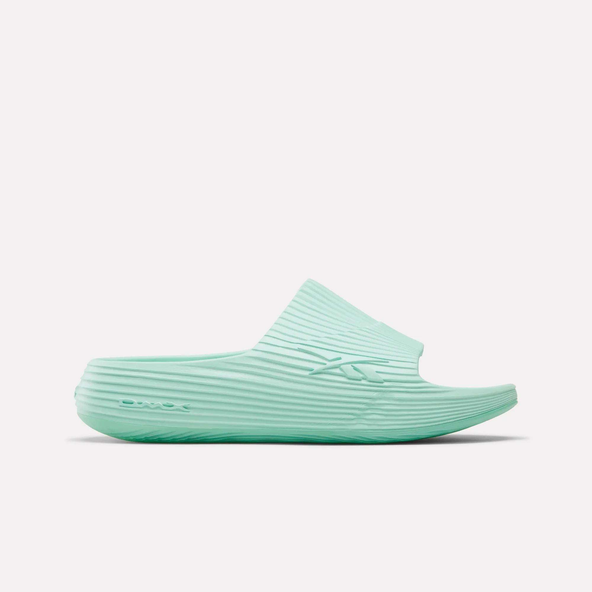 The Reebok DMX Recovery Walking Slides in light teal feature a ribbed texture, open toe, and thick sole with moving air cushioning, displayed in side profile on a white background.