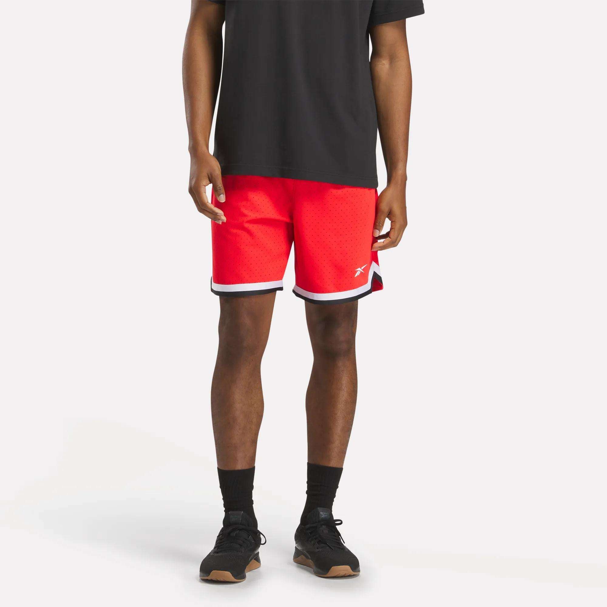A person wearing a black T-shirt, Reebok Men's Basketball Transition Shorts 7" in bright red with white trim, black socks, and black sneakers stands against a plain white background.