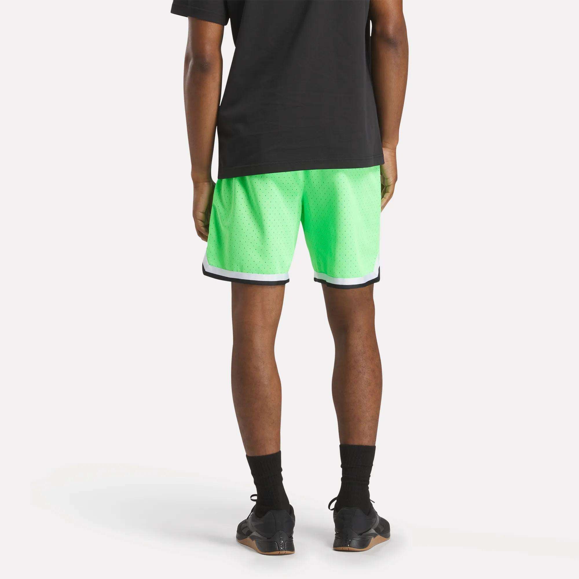 A person stands with their back to the camera wearing a black T-shirt and Reebok Men's Basketball Transition Shorts 7" in bright green with white and navy trim, paired with black socks and sneakers, against a plain white background.