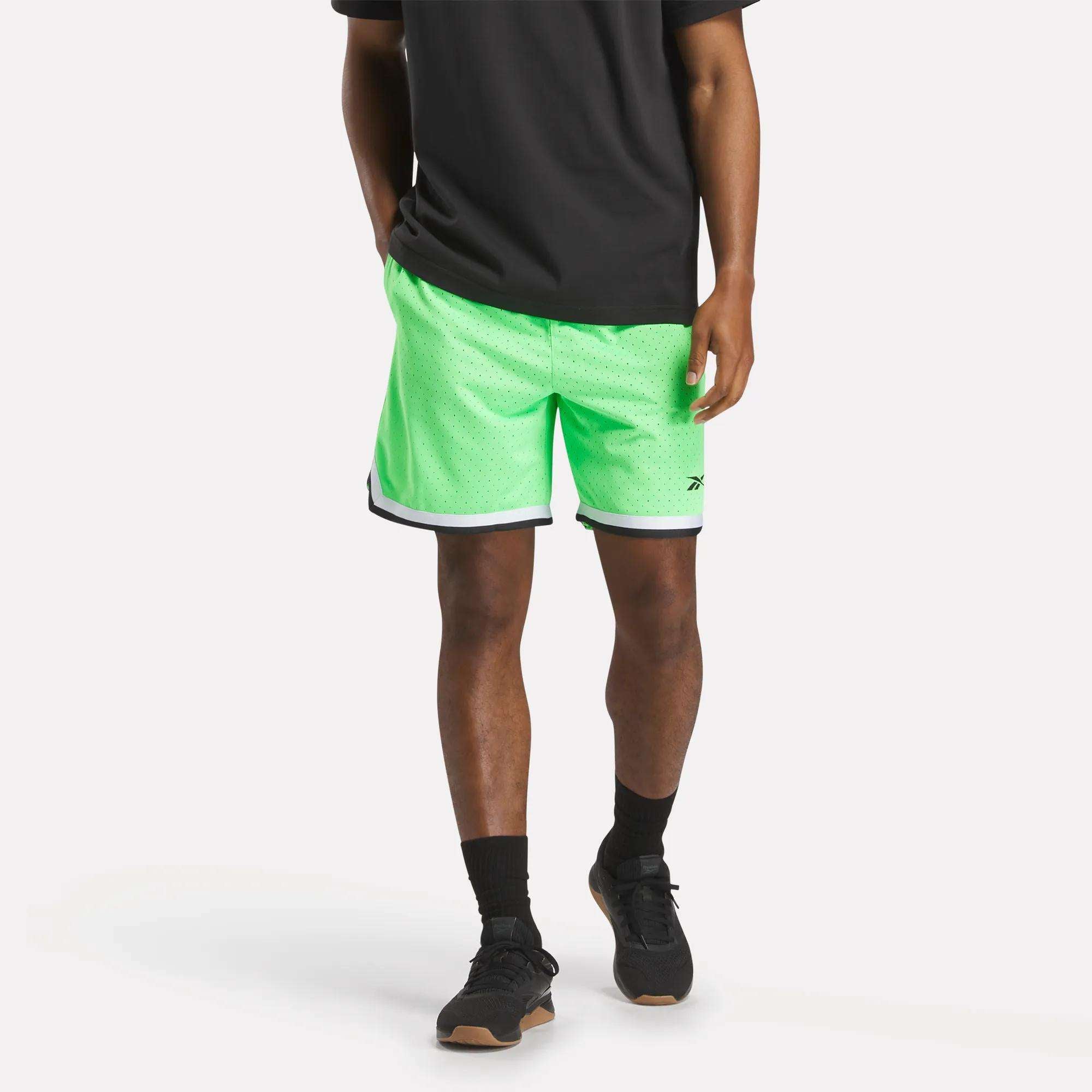 A person wearing a black T-shirt, Reebok Men's Basketball Transition Shorts 7" in neon green with black and white trim, black socks, and black sneakers stands against a plain white background.