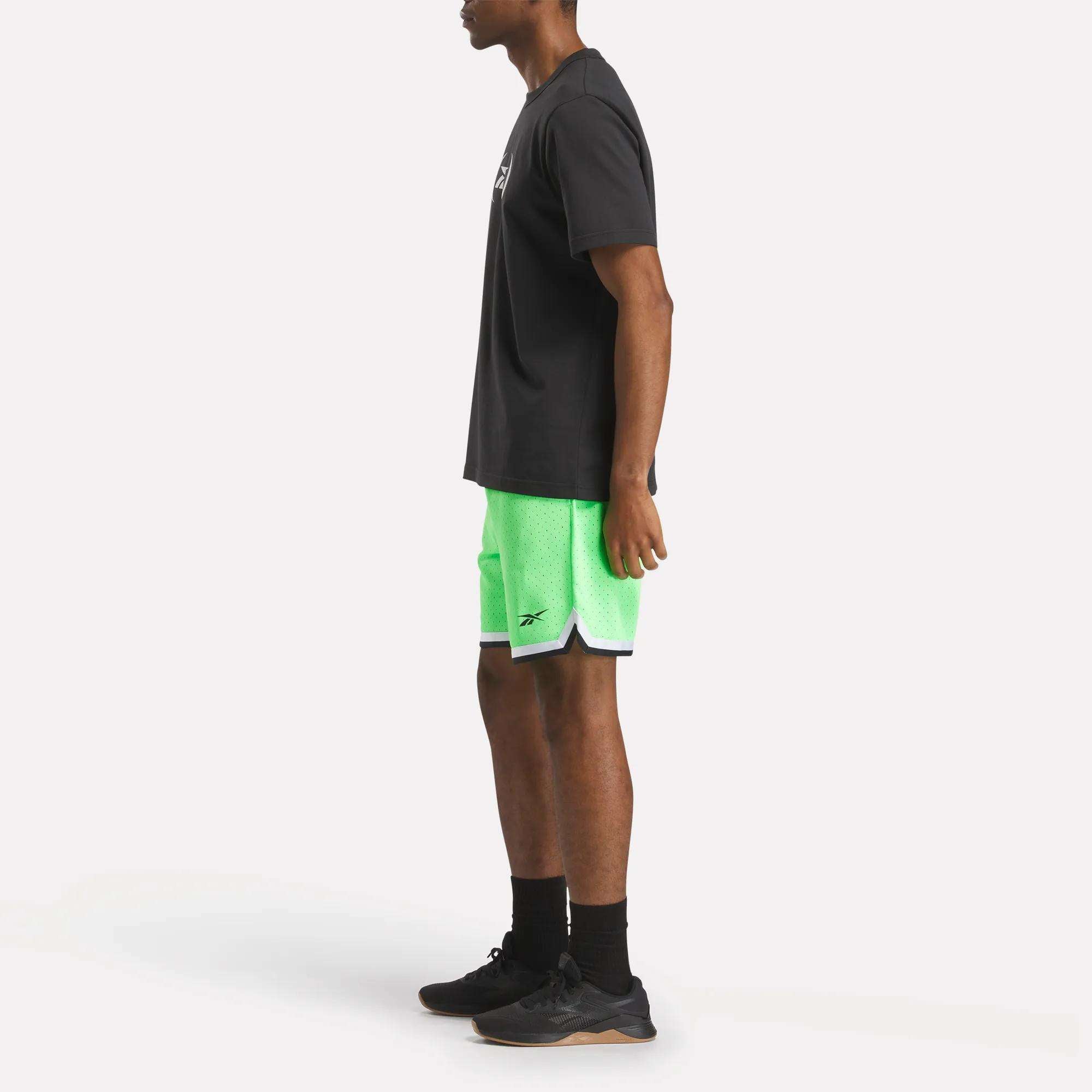 A person stands sideways wearing a black t-shirt, Reebok Men's Basketball Transition Shorts 7" in bright green with a small logo, black socks, and black sneakers against a plain white background.
