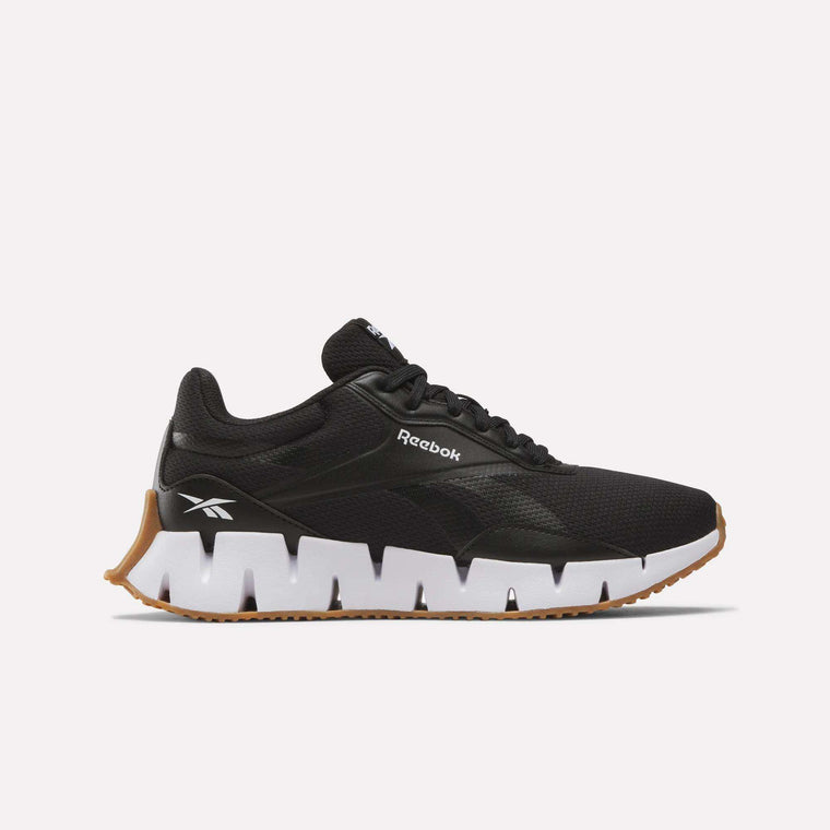 The Reebok Women's Zig Dynamica STR Shoes in black feature a white, jagged-edge innovative outsole with tan rubber and display the Reebok logo on the heel and side, shown in profile on a plain white background.
