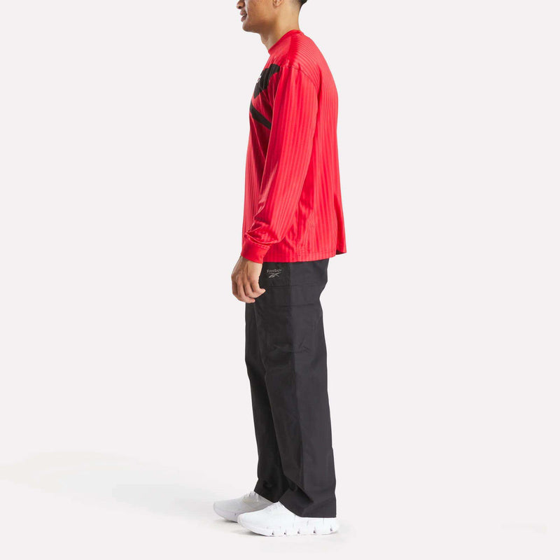 A person stands in profile against a white background, wearing the Crosscheck Long Sleeve Jersey in red with black accents, paired with black pants and white sneakers for a sporty look.