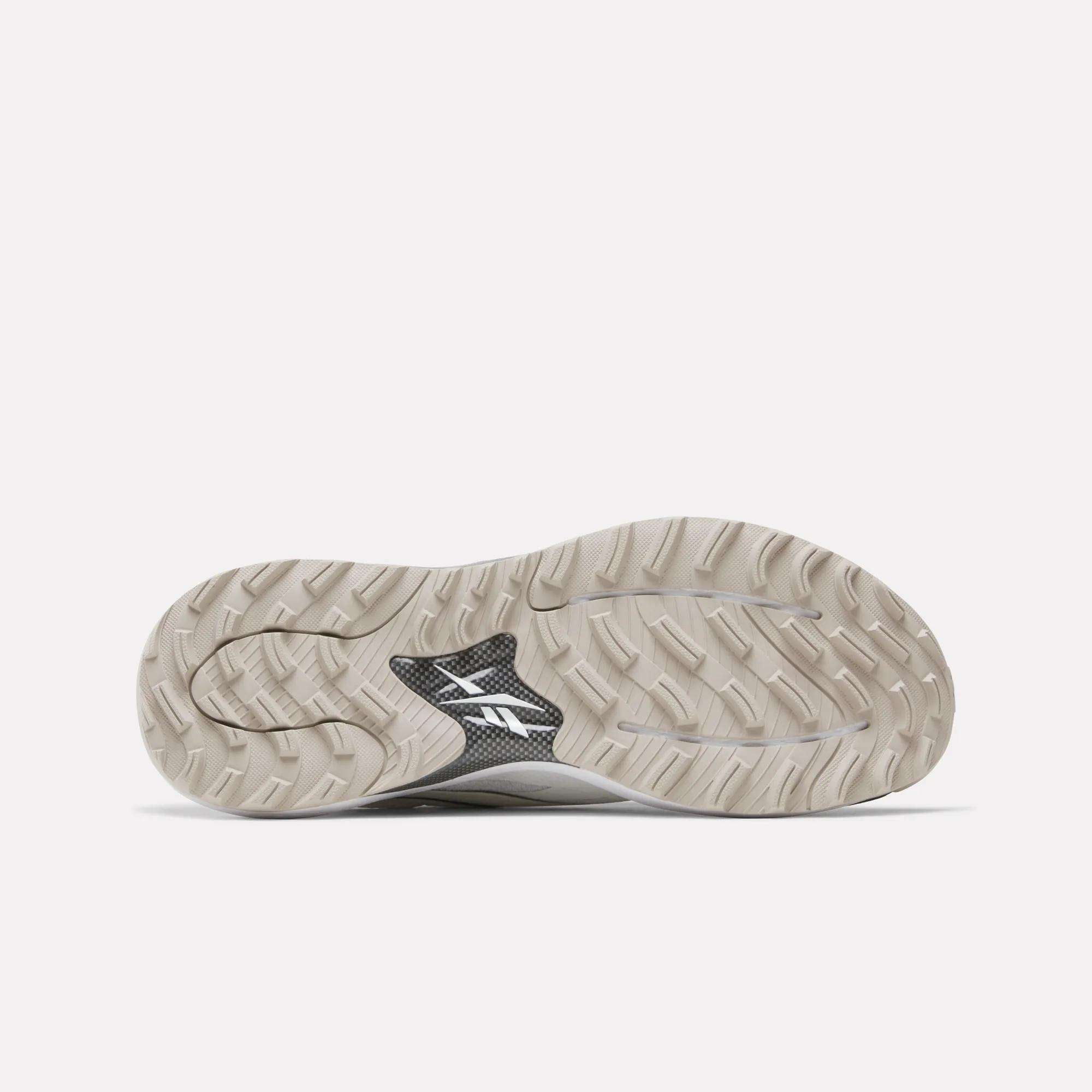 The image shows the sole of Reebok Nano Golf Shoes, featuring a beige, textured tread with a black middle section displaying a white logo—highlighting the comfort of Floatride foam. The background is plain and light-colored.
