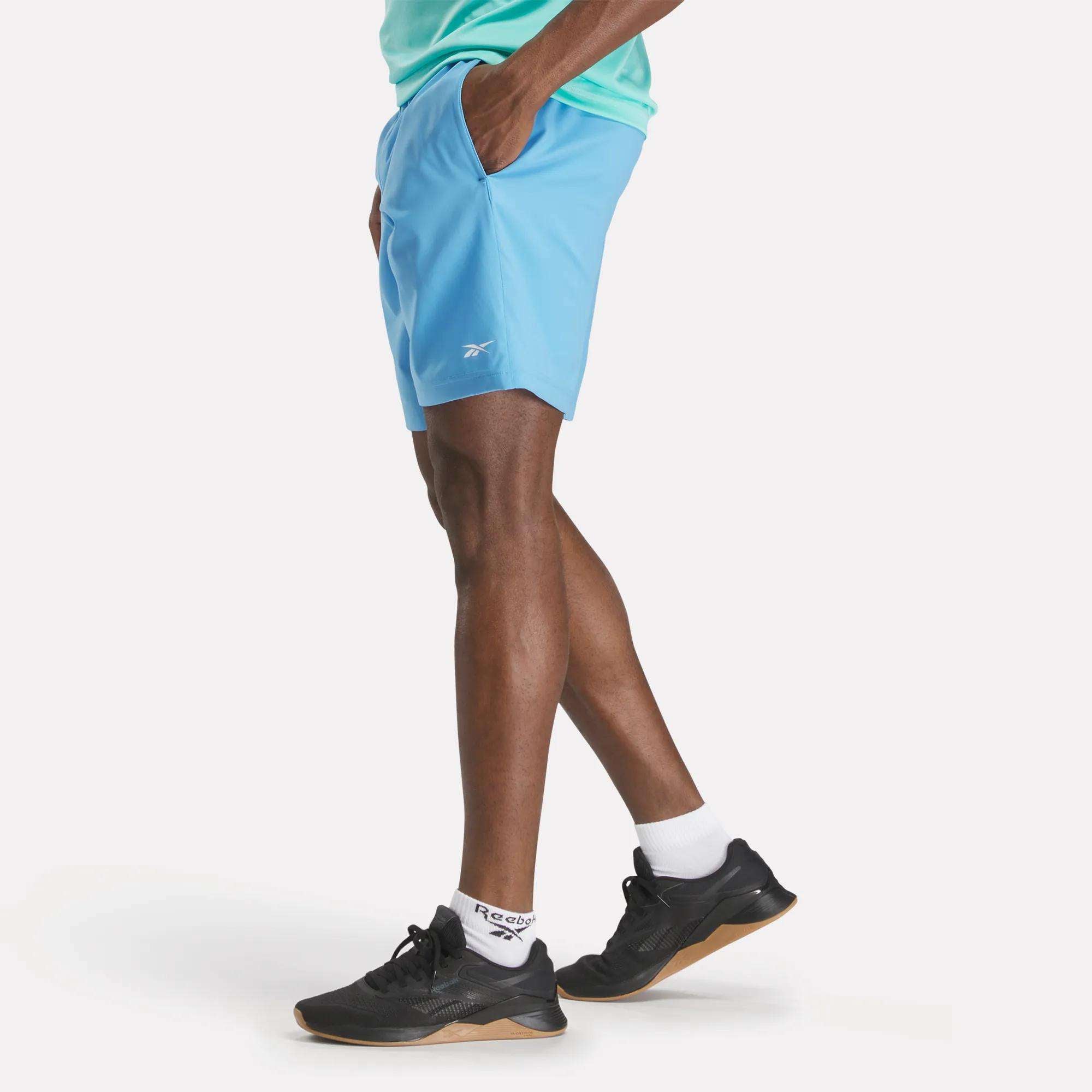 Wearing Reebok Men's Workout Ready Shorts 9" in light blue, a model stands with hands in pockets against a white background. Only the lower half of their body is visible, paired with a green shirt, white socks, and black sneakers.