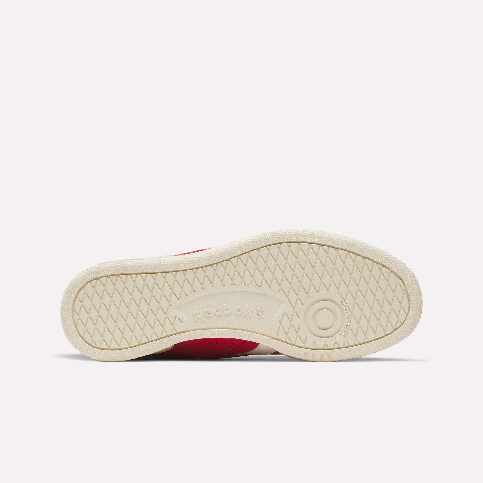 The image shows the beige sole of the Reebok Club C Ground UK Shoes with a circular pattern near the heel, textured grip, and embossed Reebok logo. The partially visible red suede upper highlights these classic sneakers.
