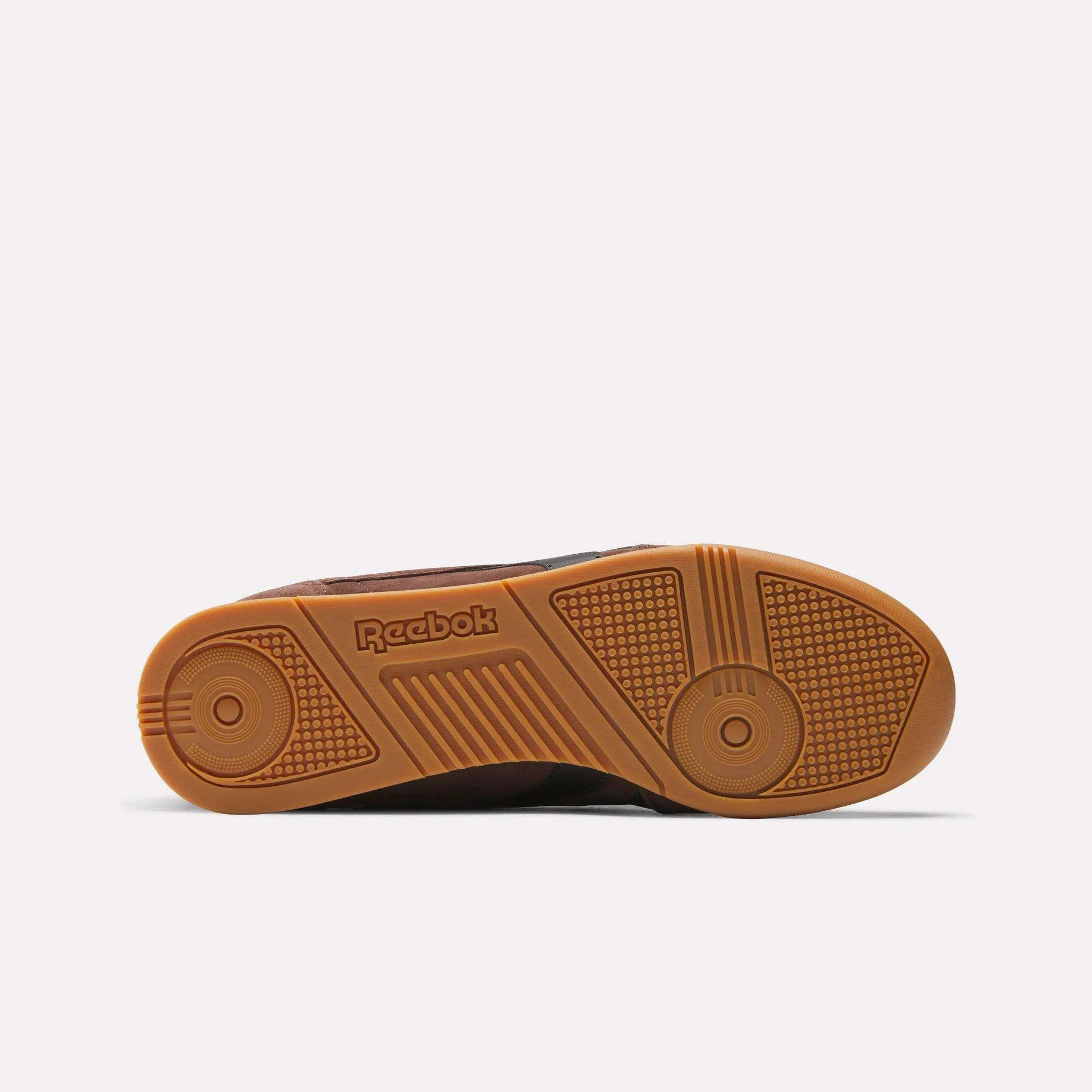 The image shows the bottom view of a brown Reebok Campio XT Shoes sole with a textured gum rubber pattern, inspired by '90s soccer style and featuring an embossed Reebok logo in the center.