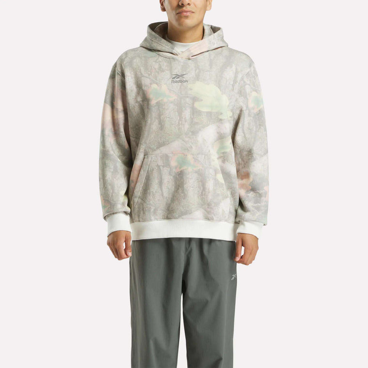 A person wearing a Reebok x MARKET Hoodie with a light camo print and hood, paired with gray pants, stands facing forward against a plain white background.